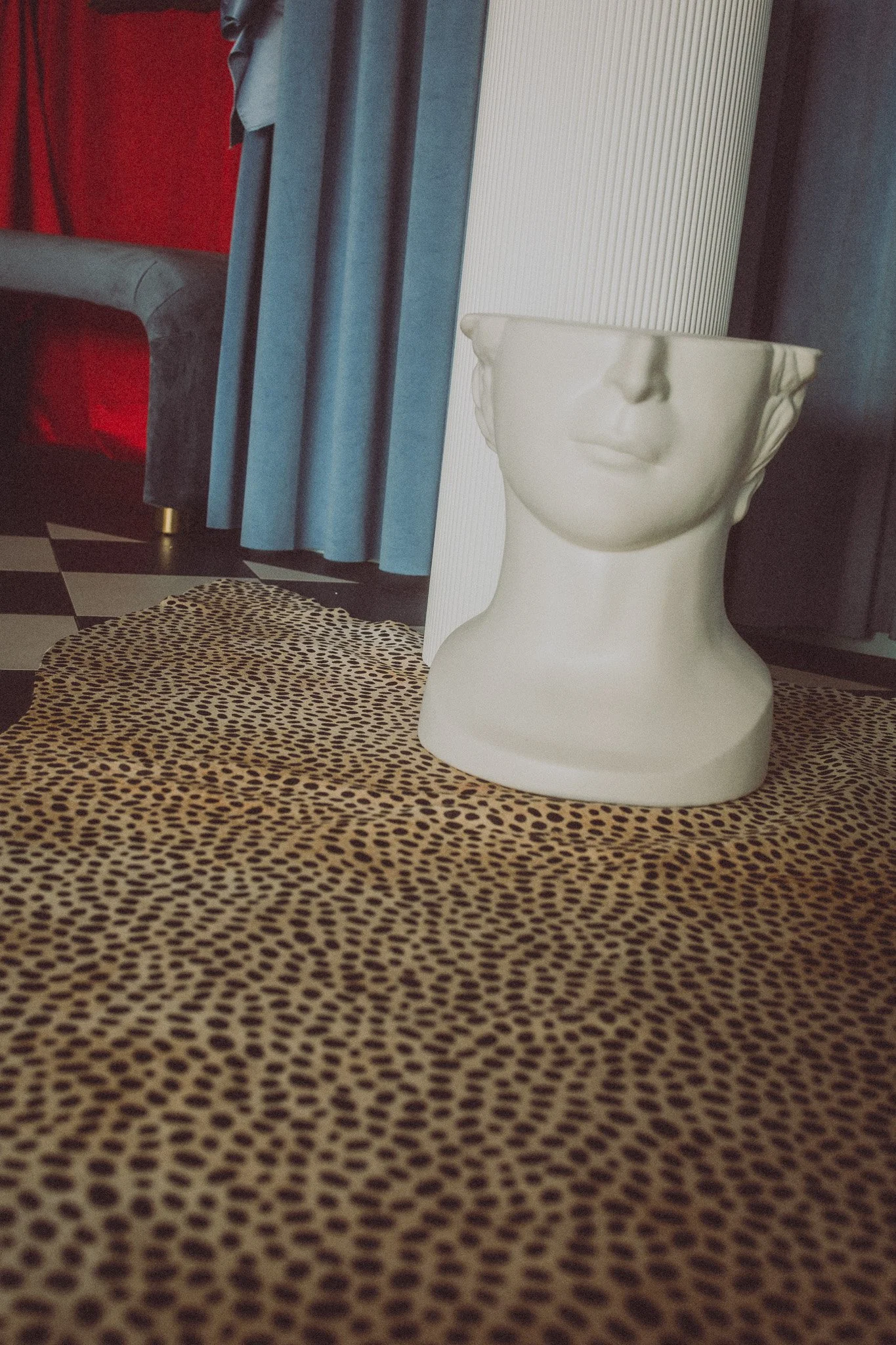 A white ceramic vase shaped like a human face, positioned on a leopard print cloth. The background includes a blue curtain, a grey sofa, and a checkered floor.