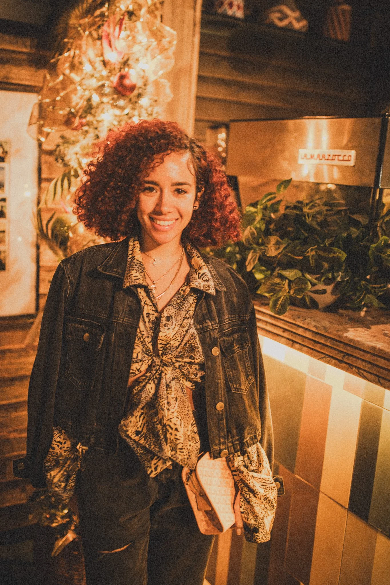 A young woman with curly hair smiling in a cozy, warmly lit indoor setting with Christmas decorations, including a decorated Christmas tree in the background, a plant, and wooden and striped decor.