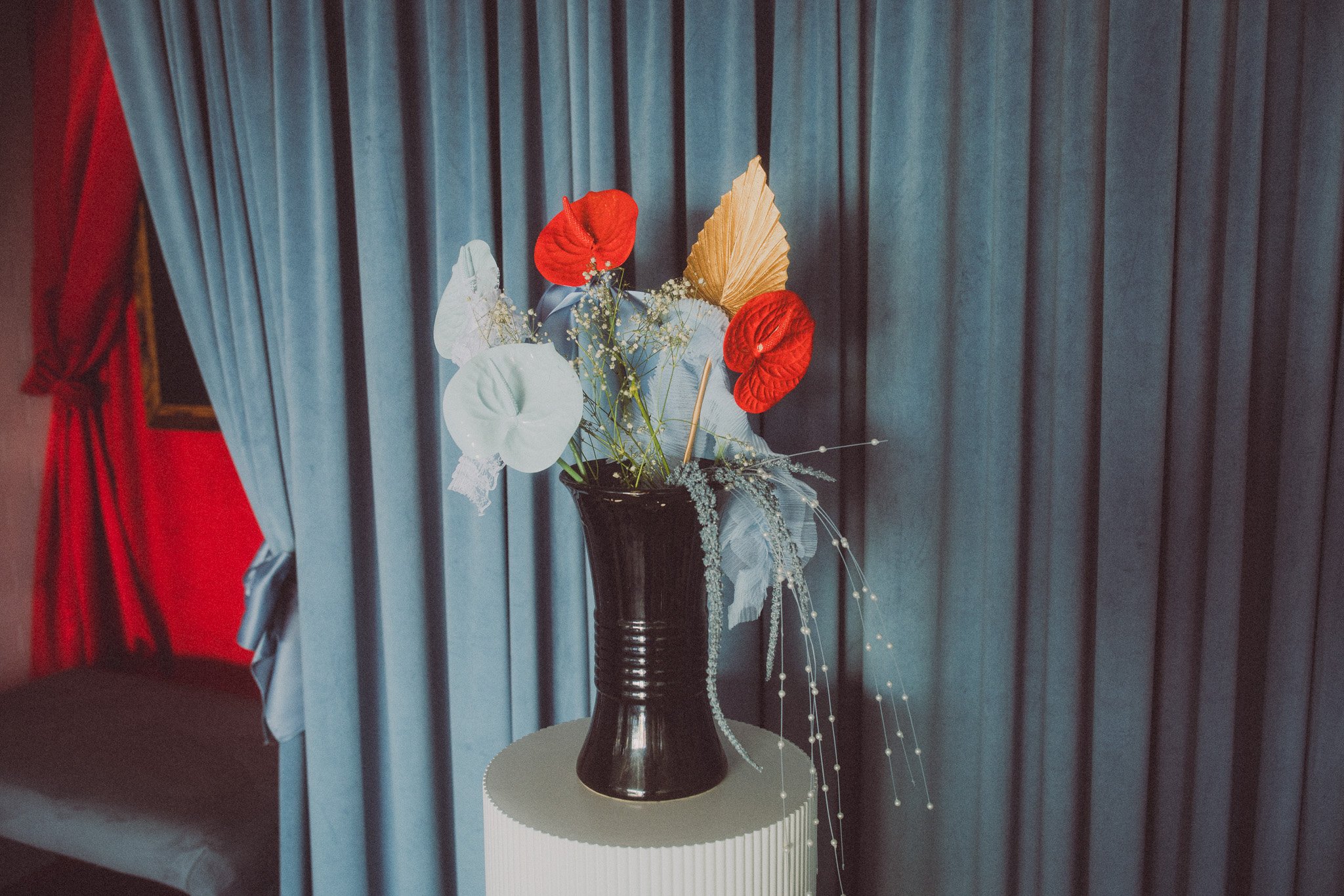 A black vase with artificial flowers, including red, white, and gold leaves, placed on a round white table in front of navy blue curtains.