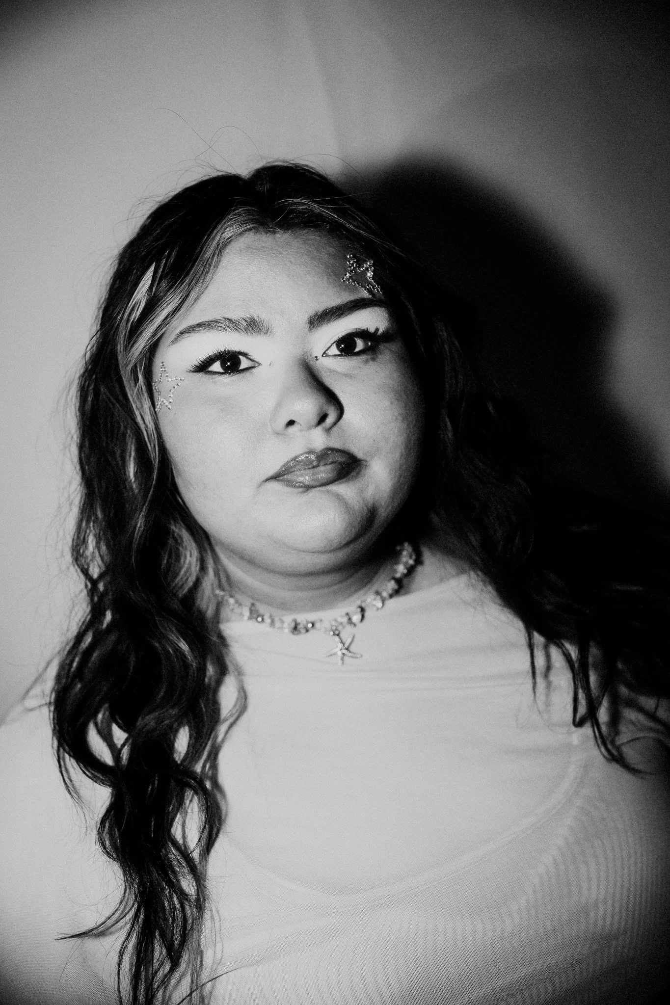 Black and white portrait of a woman with long, wavy hair, decorated with star-shaped face stickers, wearing makeup and a necklace, looking at the camera.