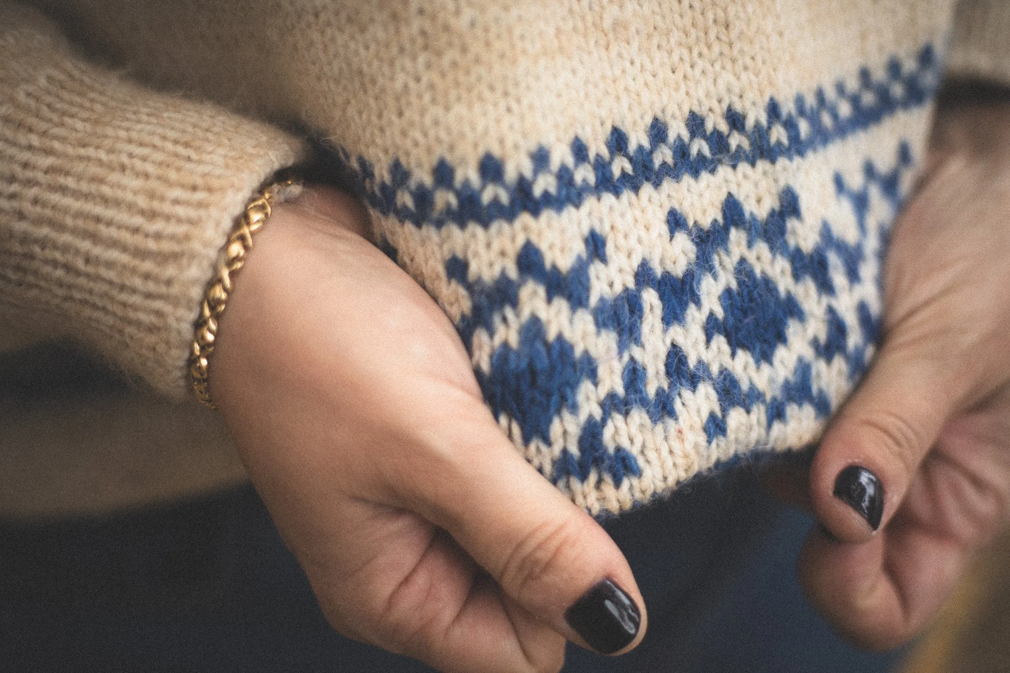 Close-up of a person's hand with black nail polish holding a knitted beige sweater with blue snowflake patterns.