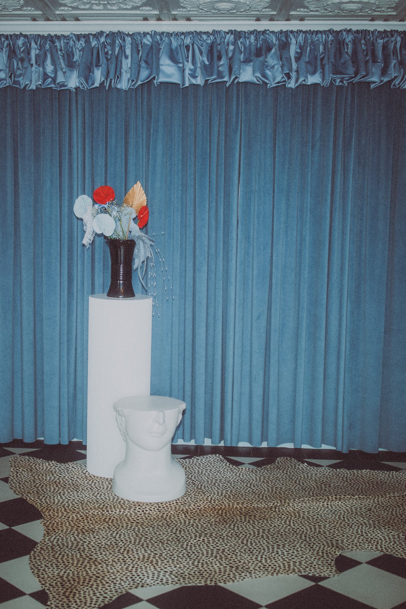 A decorative setup with a blue curtain background, a white pedestal with a black vase containing artificial flowers, and a white bust-shaped object on the floor in front of the pedestal. The floor has a patterned carpet and black and white checkered 