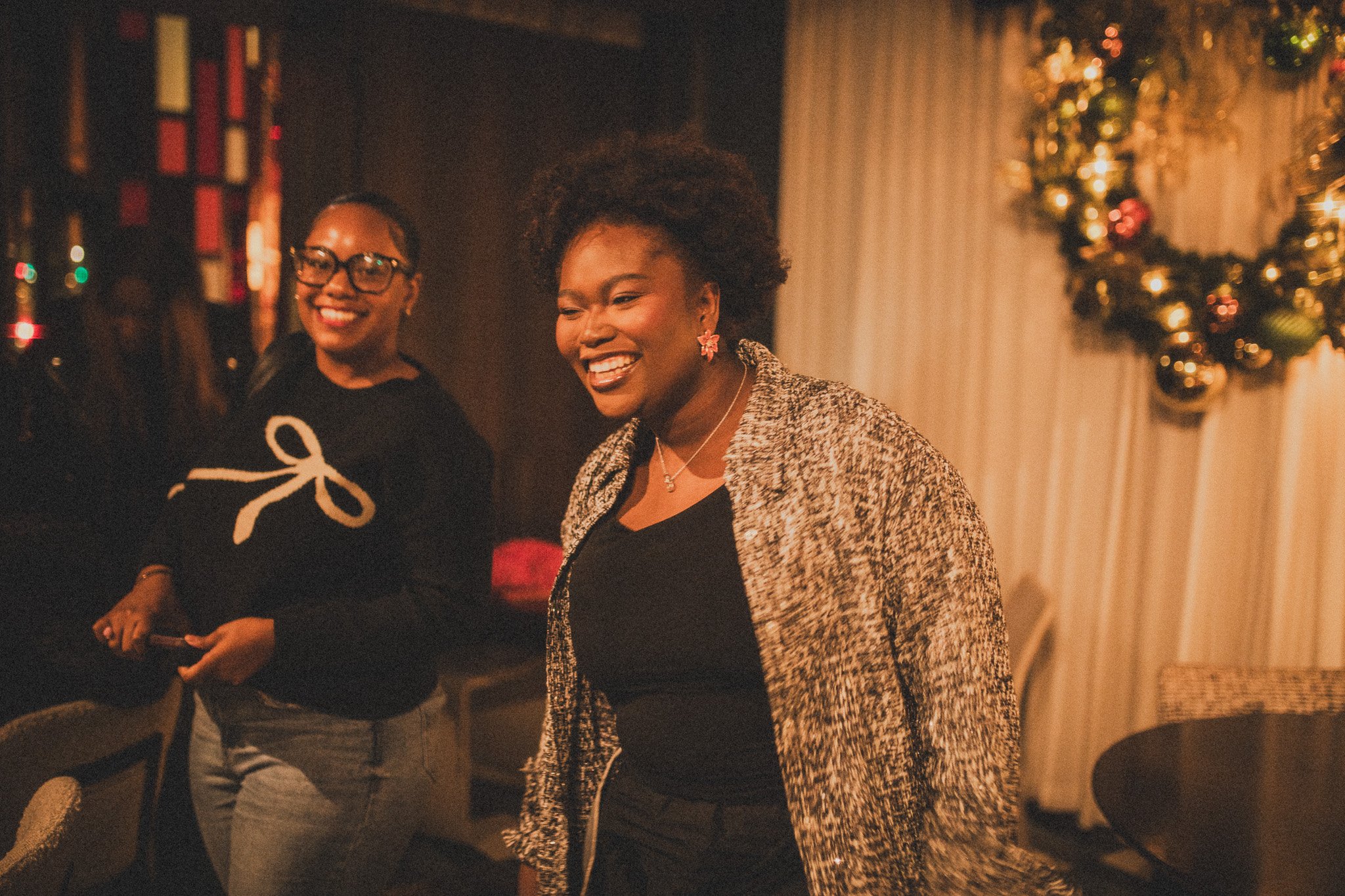 Two women smiling and laughing at a festive gathering, decorated with Christmas ornaments and a wreath, in a cozy indoor setting.