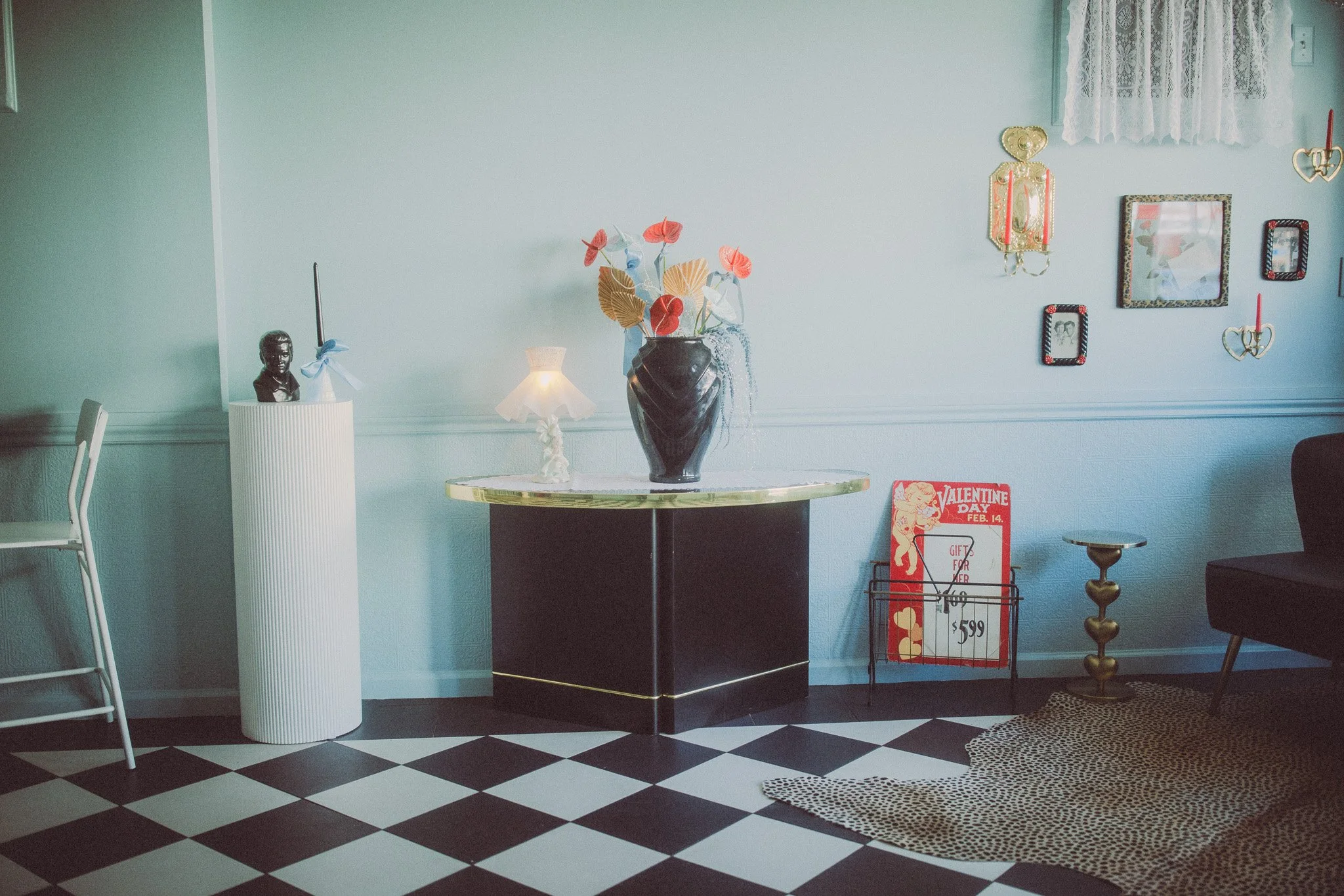 Living room with a black and white checkered floor, white walls, and various decorations including a large black vase with artificial flowers on a table in the center, a small white lamp, a gold-colored side table, and a wall with picture frames and 