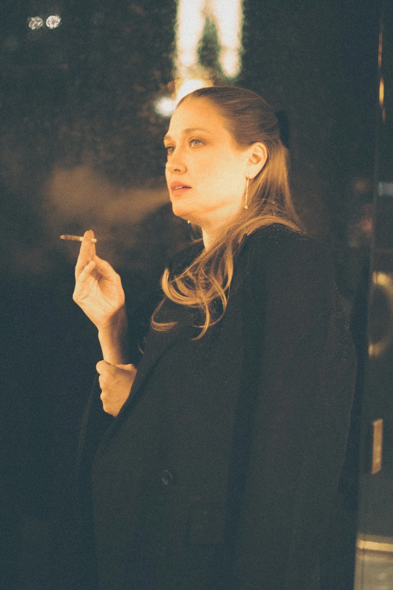 A woman with long, wavy blonde hair and light skin is holding a cigarette in her right hand. She is wearing a black blazer and earrings, and is standing indoors with a blurry background.
