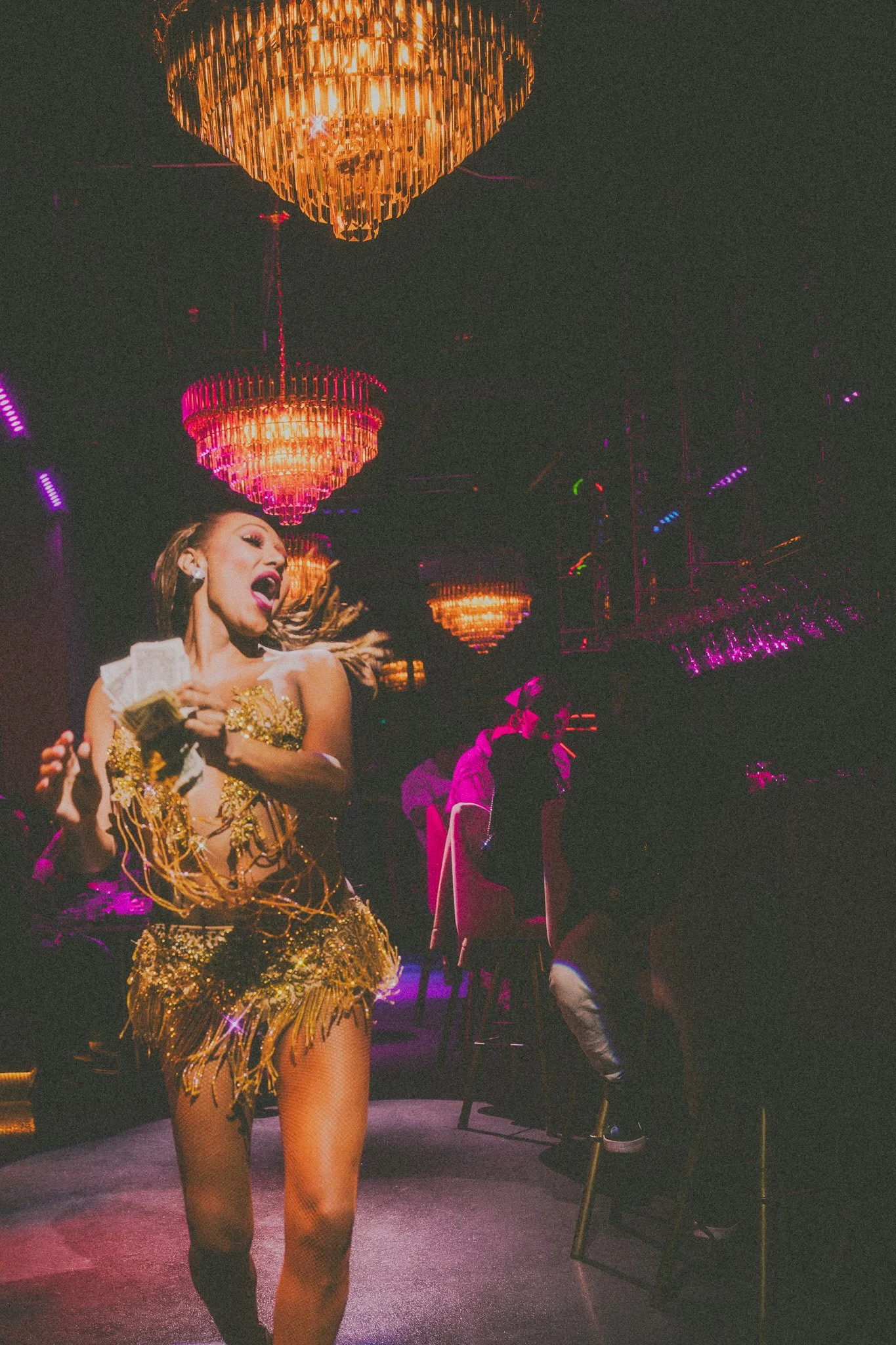 A woman dressed in an elaborate gold costume is singing passionately on stage under large, glowing chandeliers in a dark, vibrant nightclub with neon purple lighting.
