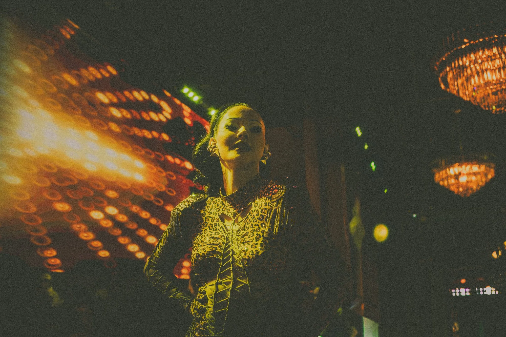 A woman with dark hair, wearing a leopard print outfit and statement earrings, stands in a dimly lit room with colorful, blurred lights and chandeliers overhead.