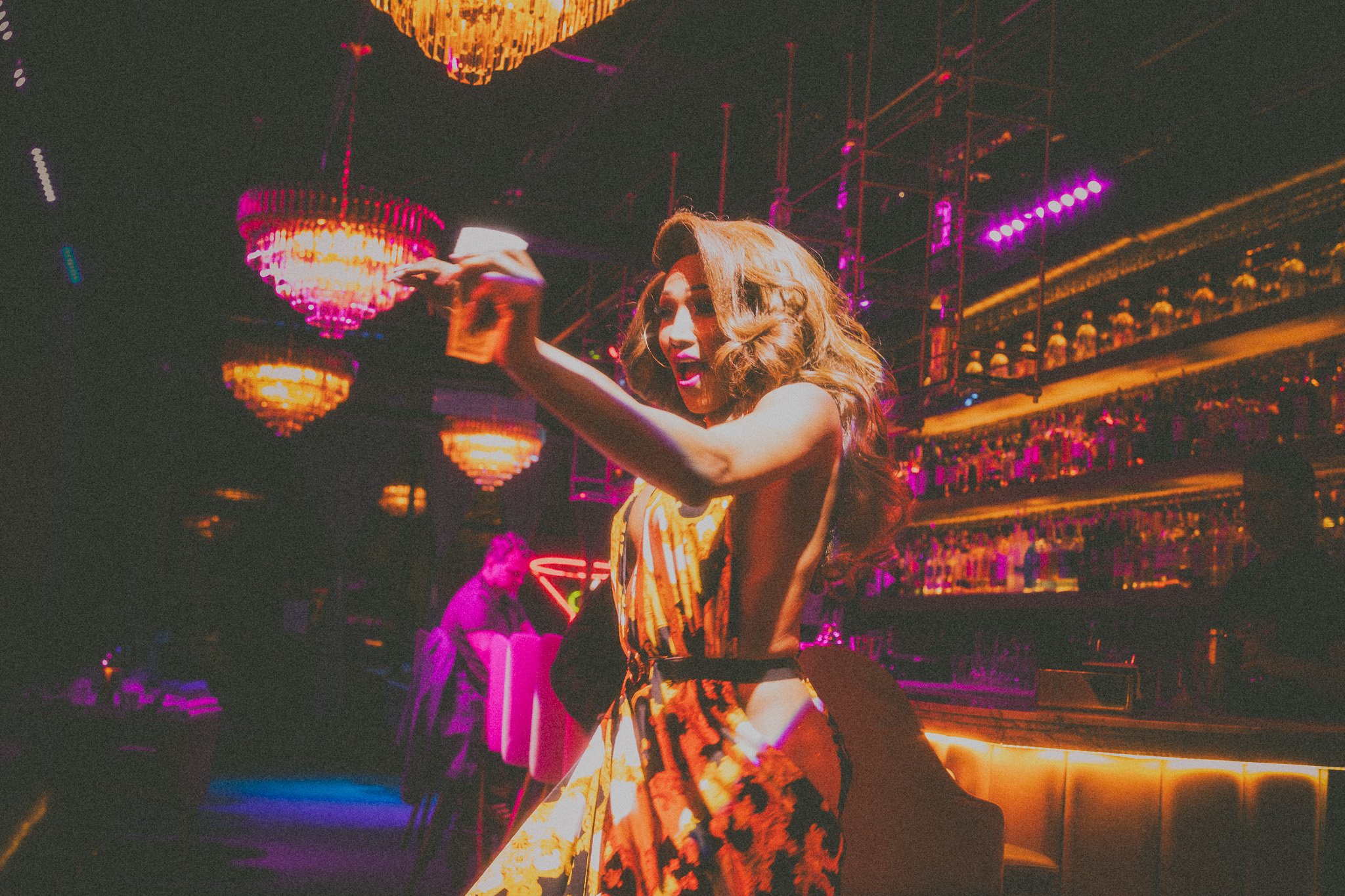 A woman in a colorful dress dancing or posing in a dimly lit nightclub with hanging chandeliers and a bar in the background.