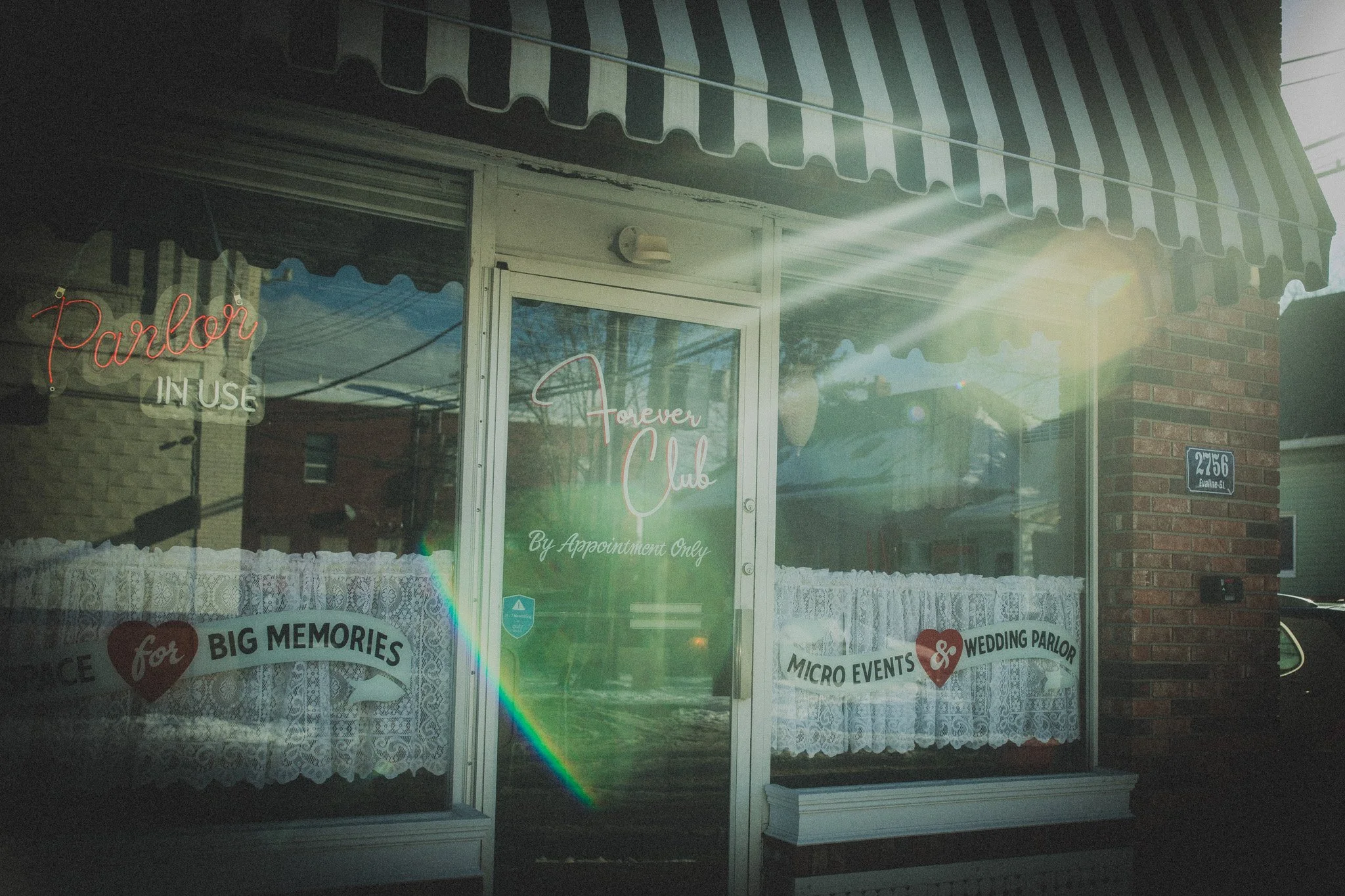 The storefront of a wedding parlor with glass windows, lace curtains, and neon signs. The signs read "Parlor in Use" and "Forever Club by Appointment Only." There are decorative signs on the window with words "for big memories" and references to micr