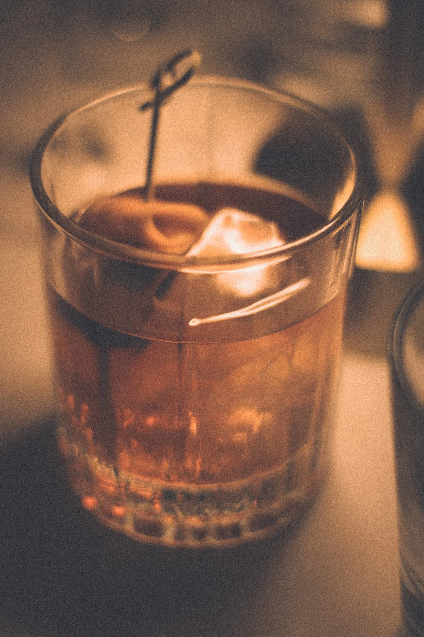 A close-up of a glass with a dark drink and ice cubes, garnished with a cherry and an orange slice, under warm lighting.