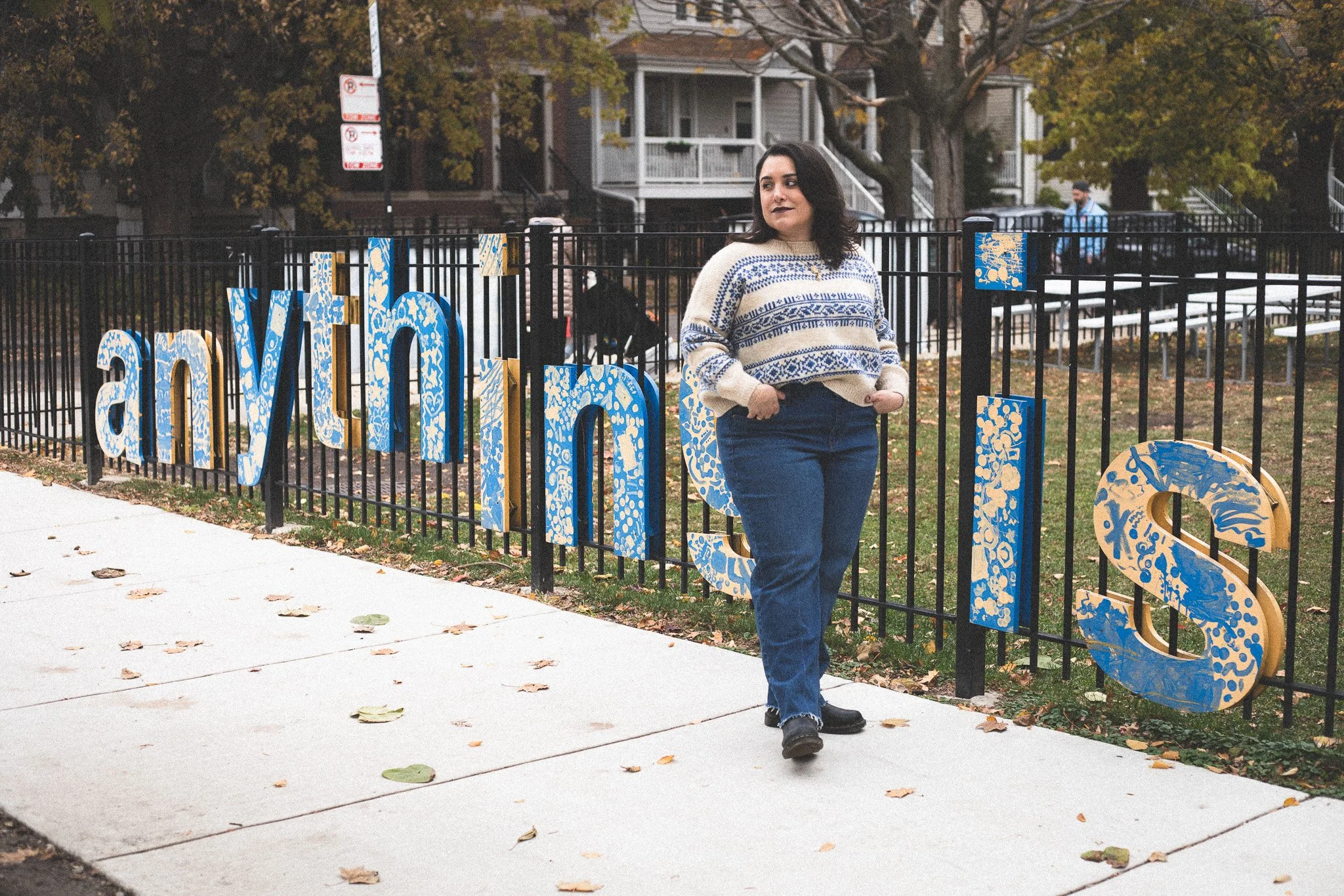 A woman with dark hair and dark lipstick stands on a sidewalk next to a colorful fence with large decorative blue and yellow letters that spell out 'anywhere' on a fall day.