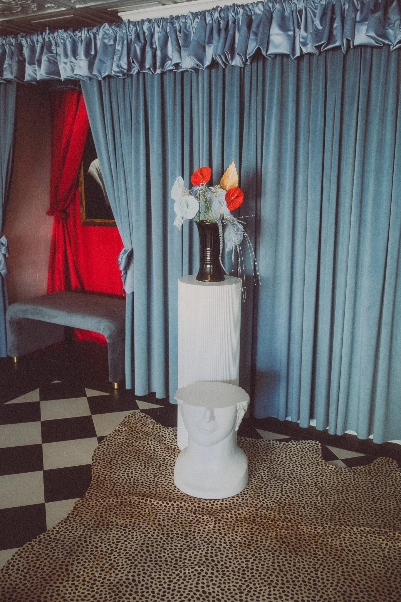 A room decorated with blue curtains, a black vase with red and white flowers, a white pedestal with a sculpture of a human bust, and a leopard print rug on a checkered black-and-white floor.