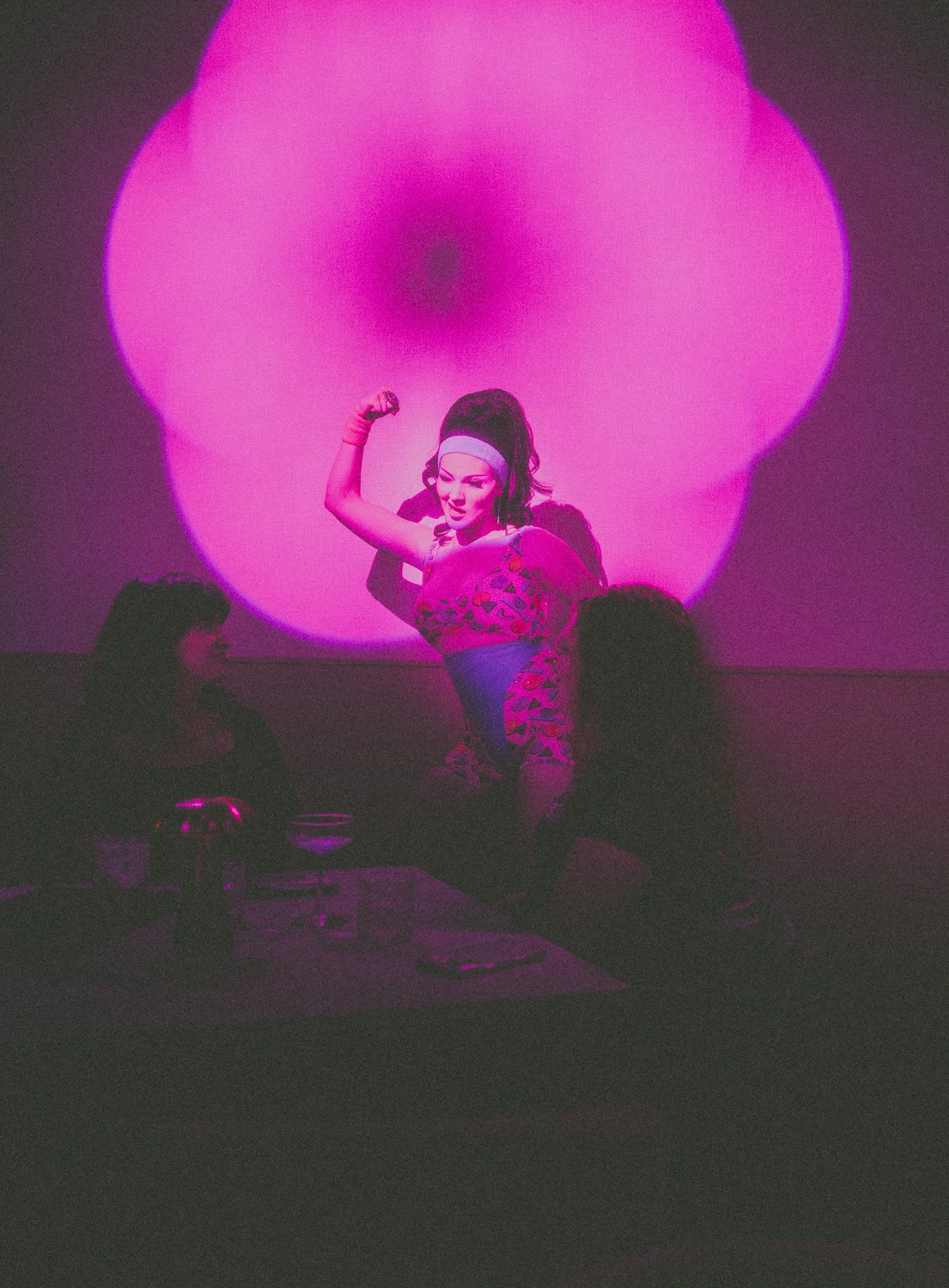 A woman in a pink and purple athletic outfit with a headband flexes her arm muscle in front of a large pink circular light projection. Two women sit on a couch watching her, with snacks and drinks on a table nearby, in a dimly lit room.