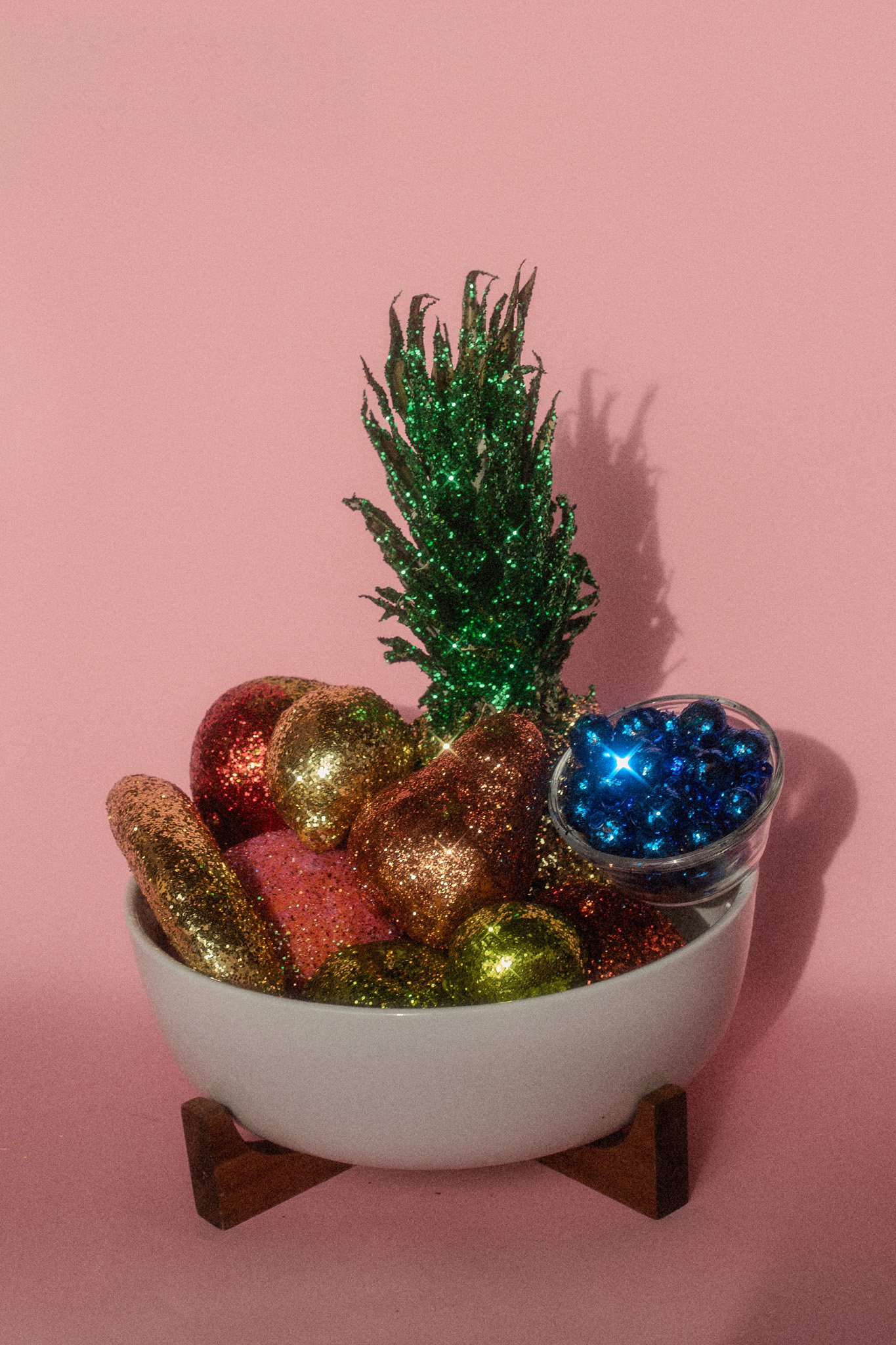 Decorative bowl filled with glittery, colorful chocolate candies, a small green glittery plant, and a glass container of blue, shiny ornaments, all on a white dish stand against a pink background.