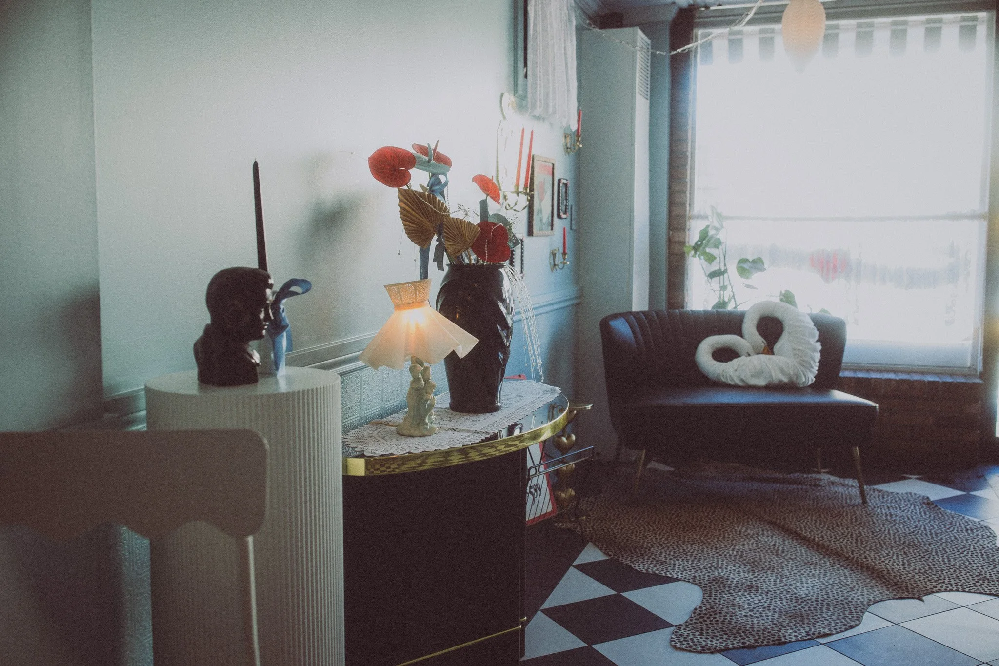 A cozy living room with a black armchair, a large window with blinds, plants, and a leopard-print rug. Decor includes a black vase with paper flowers, a small lamp, a bust sculpture, and wall-mounted candles.