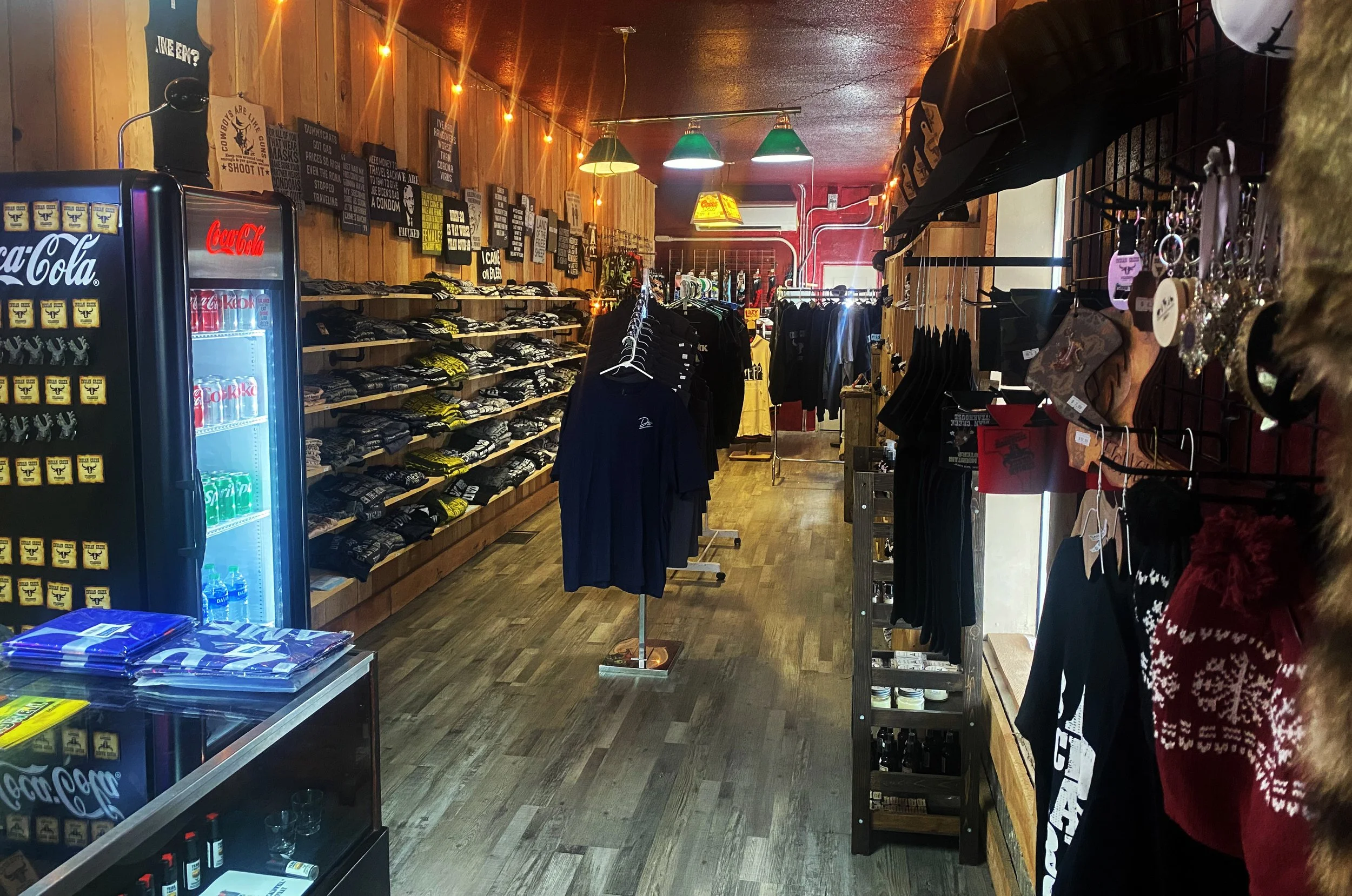 Inside the retail store with wooden walls, clothing racks with shirts, hoodies, and jackets, shelves with folded clothes, a Coca-Cola refrigerator, and decorative lights hanging from the ceiling.