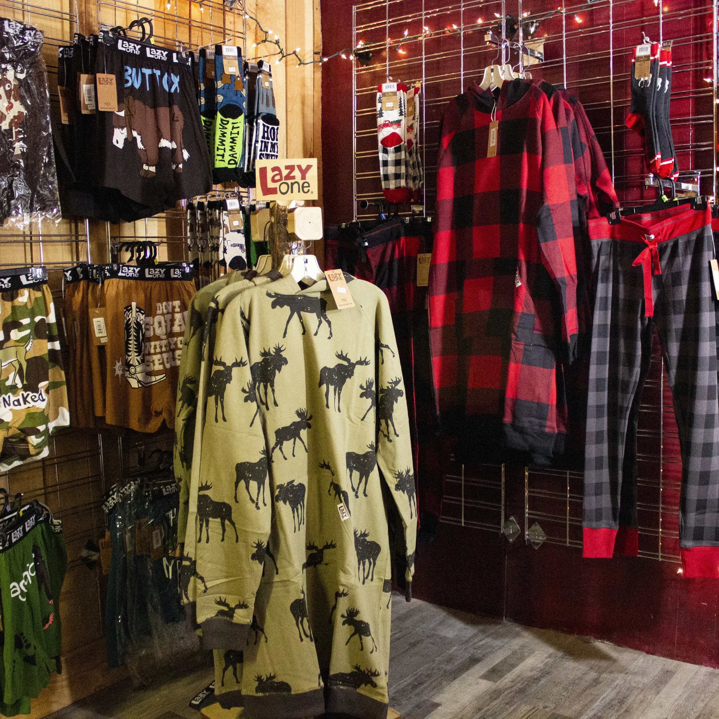 Clothing display in  store featuring pajamas and loungewear. The display includes moose and elk prints, a red and black checkered flannel, and matching pajama pants with checkered patterns. Christmas string lights are hung in the background.