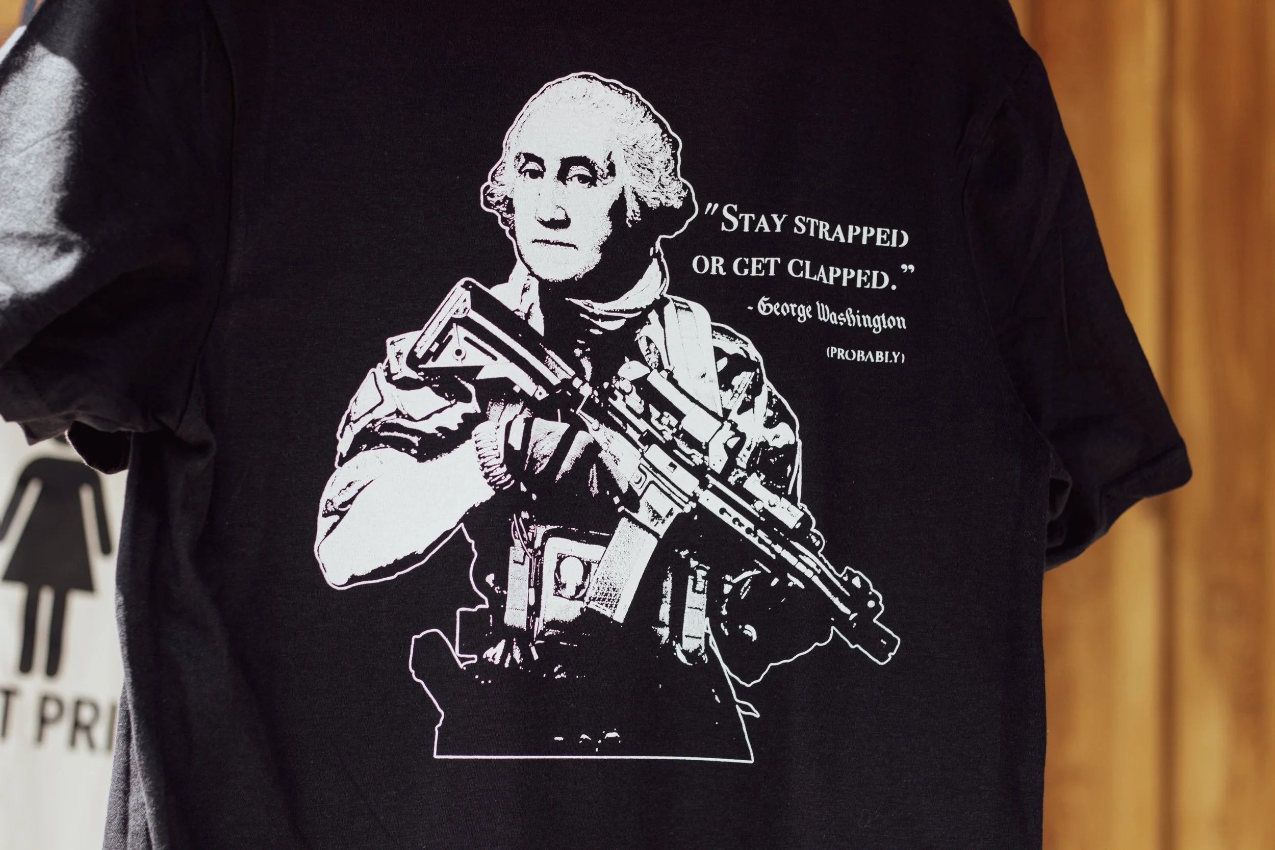 Black T-shirt with a white illustration of George Washington holding a firearm and a quote that says 'Stay strapped or get clapped,' attributed to George Washington, with the note 'probably'