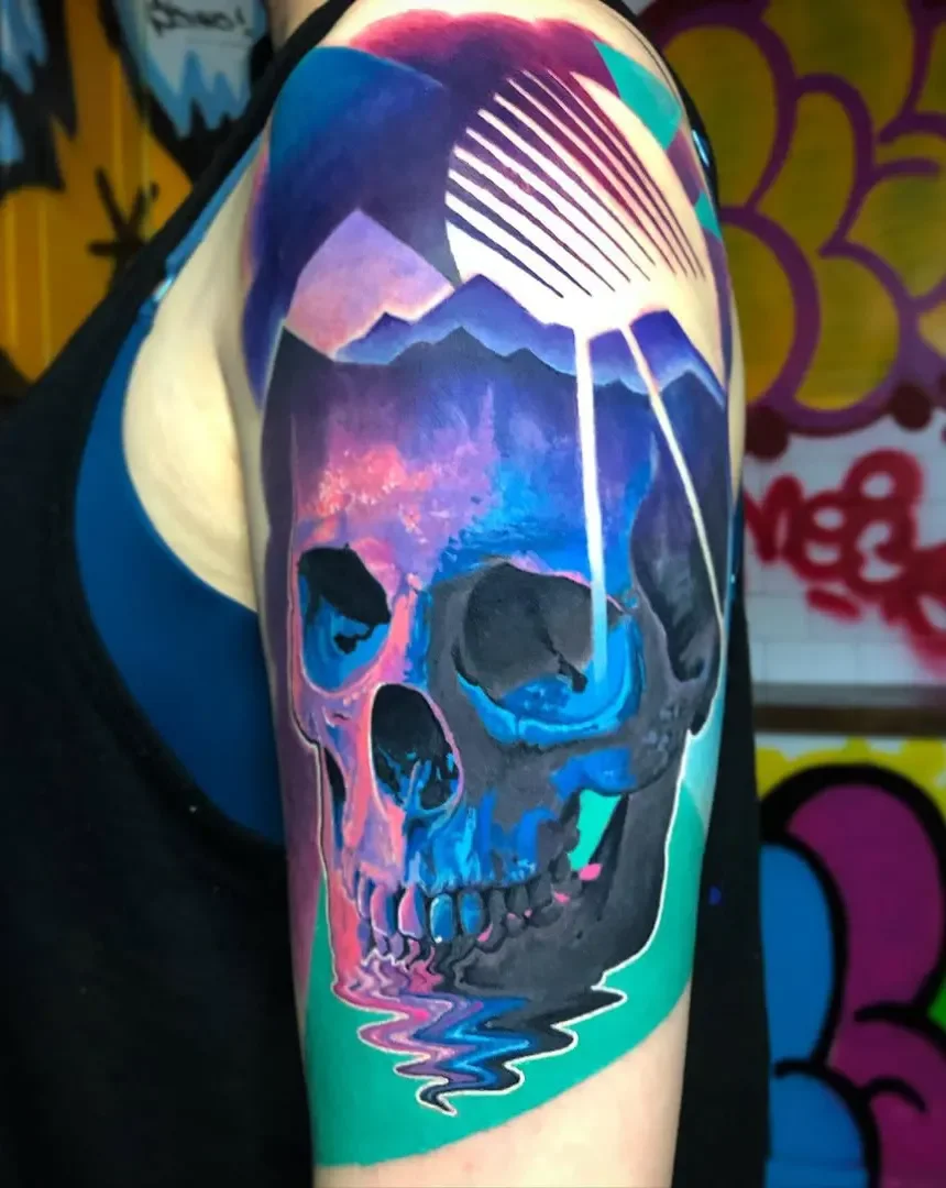 Colorful tattoo of a skull with a sunset, mountains, and a rainbow on a person's upper arm.