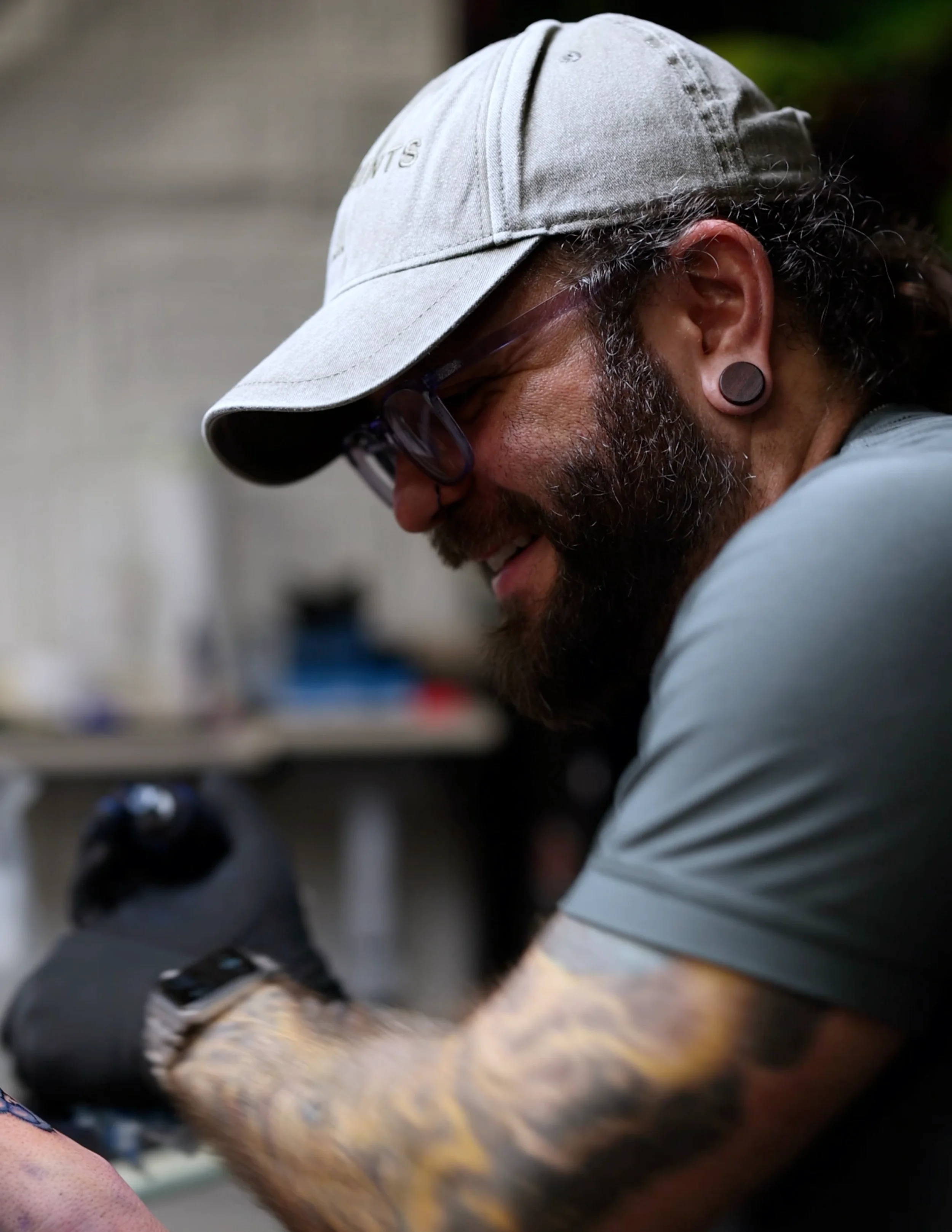 A man with a beard, wearing a gray cap, glasses, and a gray shirt, is smiling while tattooing.