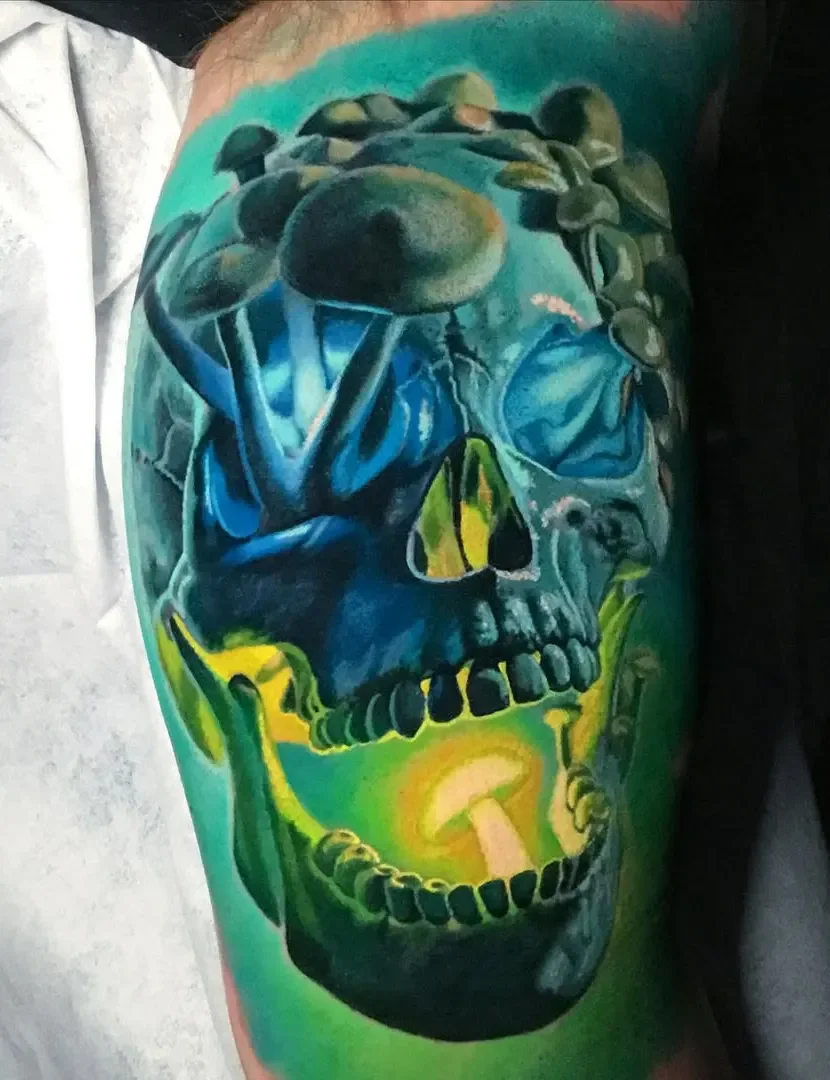 Colorful tattoo of a human skull on a person's arm, with mushrooms growing on top of the skull, and vibrant shades of blue, green, yellow, and black.