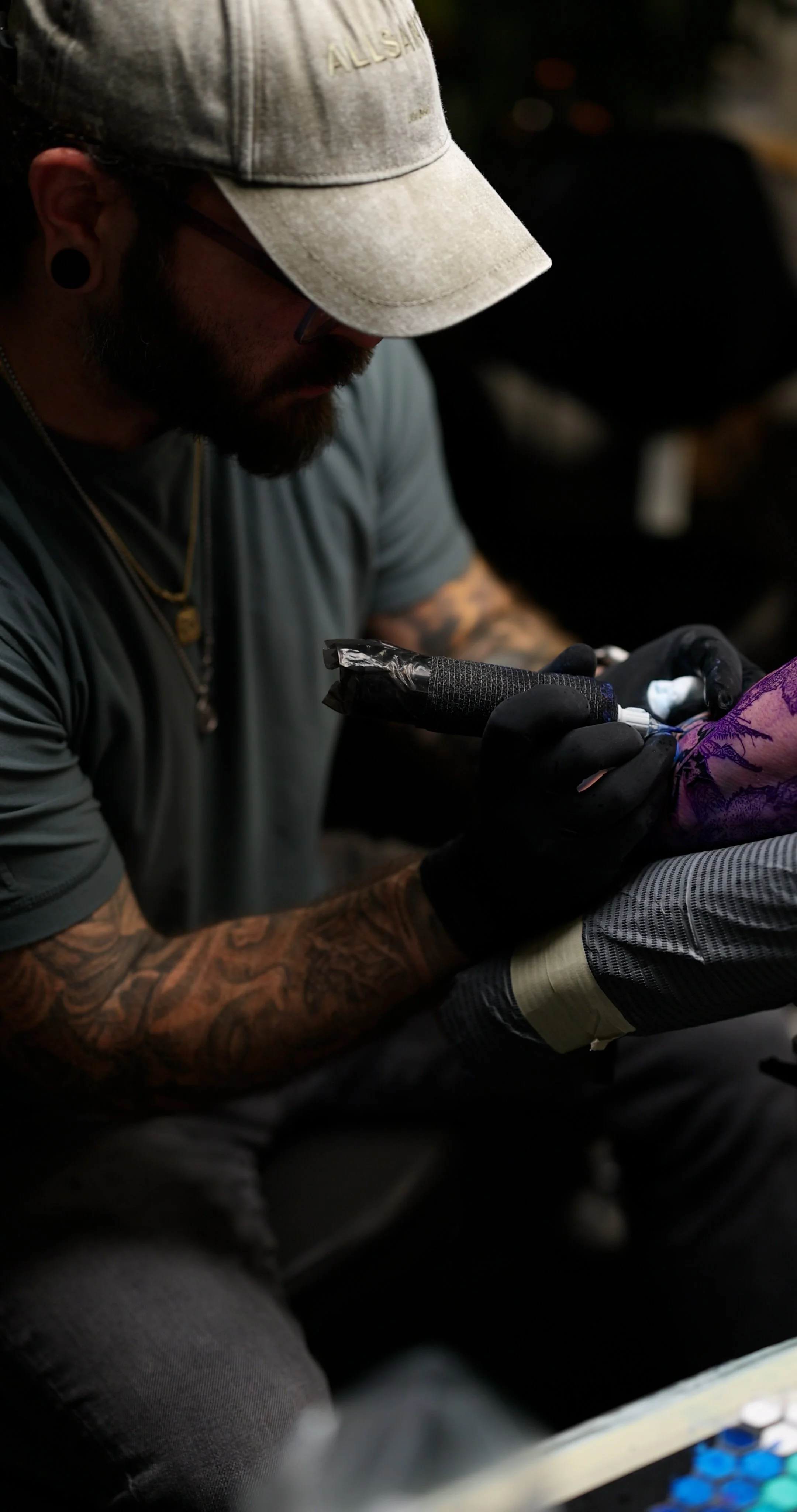 A tattooed man wearing a beige cap, glasses, and black gloves is drawing or tattooing on a person's skin with a tattoo machine.