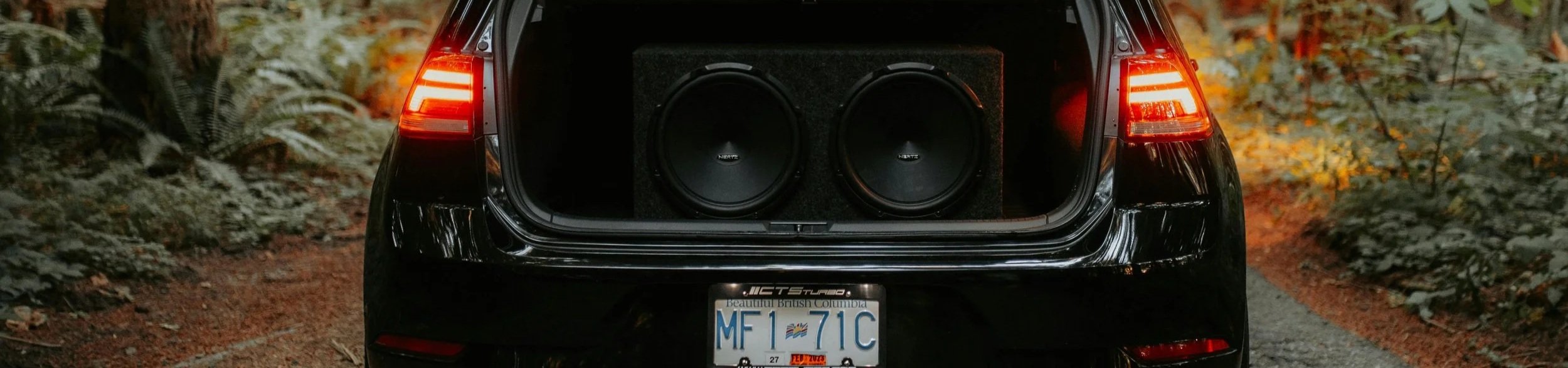 The rear of a black car with the trunk open, housing a large speaker system with two subwoofers, on a wooded dirt path during autumn.