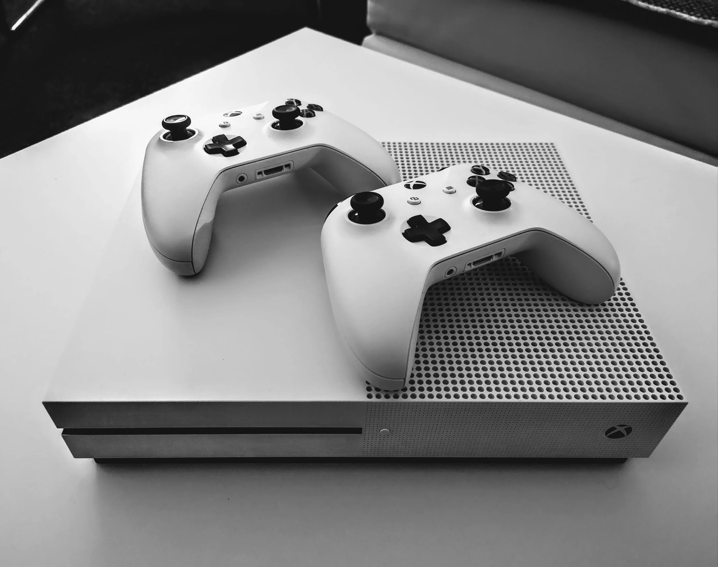 A white Xbox Series X gaming console with two white controllers placed on top, on a white surface with a perforated pattern and a dark background.