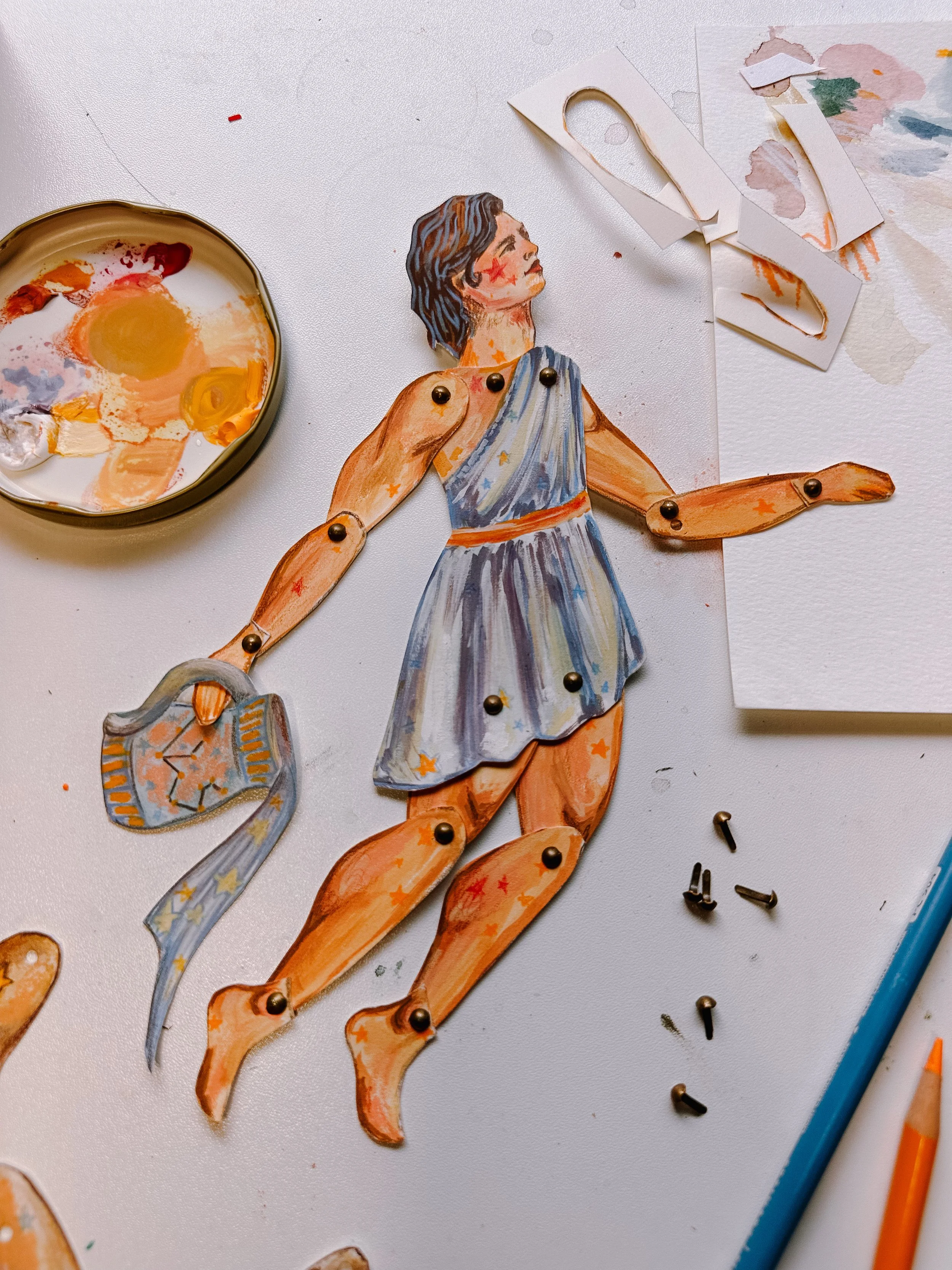 Artistic paper doll of a woman in a blue dress with painted details, attached with small metallic pins, on a white surface with painting supplies and cut-out paper pieces nearby.