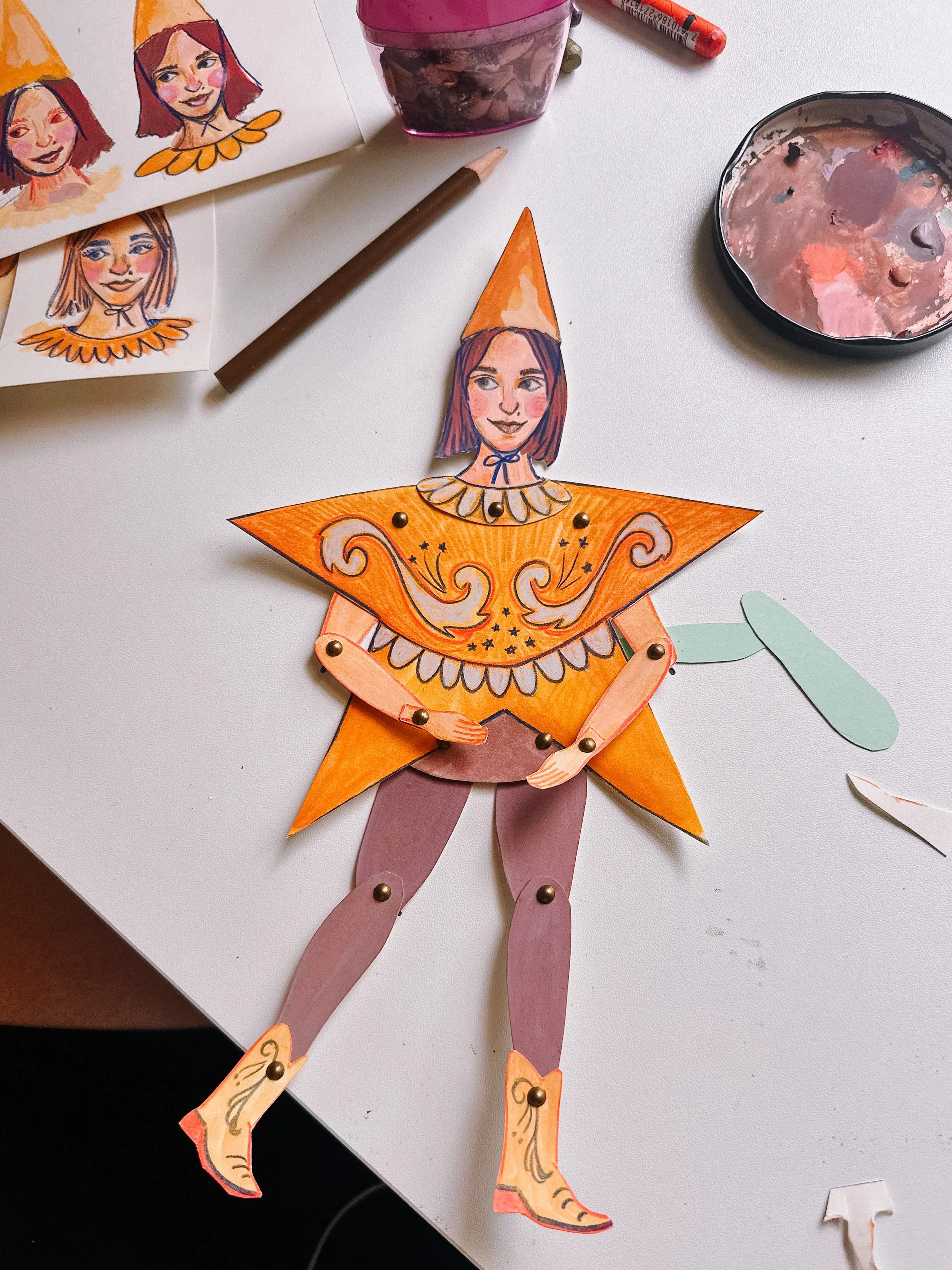 A handcrafted paper doll dressed as a clown, with a face illustration, orange star-shaped body, and movable limbs, surrounded by art supplies and sketches on a white desk.