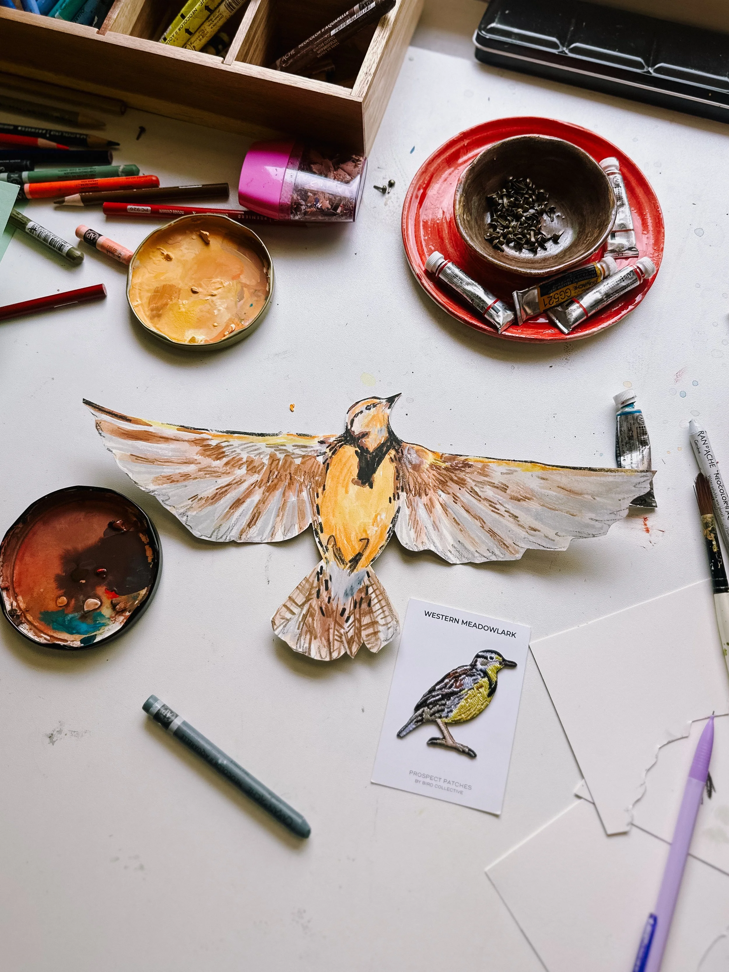 A handmade illustration of a bird with outstretched wings on a white table, surrounded by watercolor paints, brushes, and art supplies.
