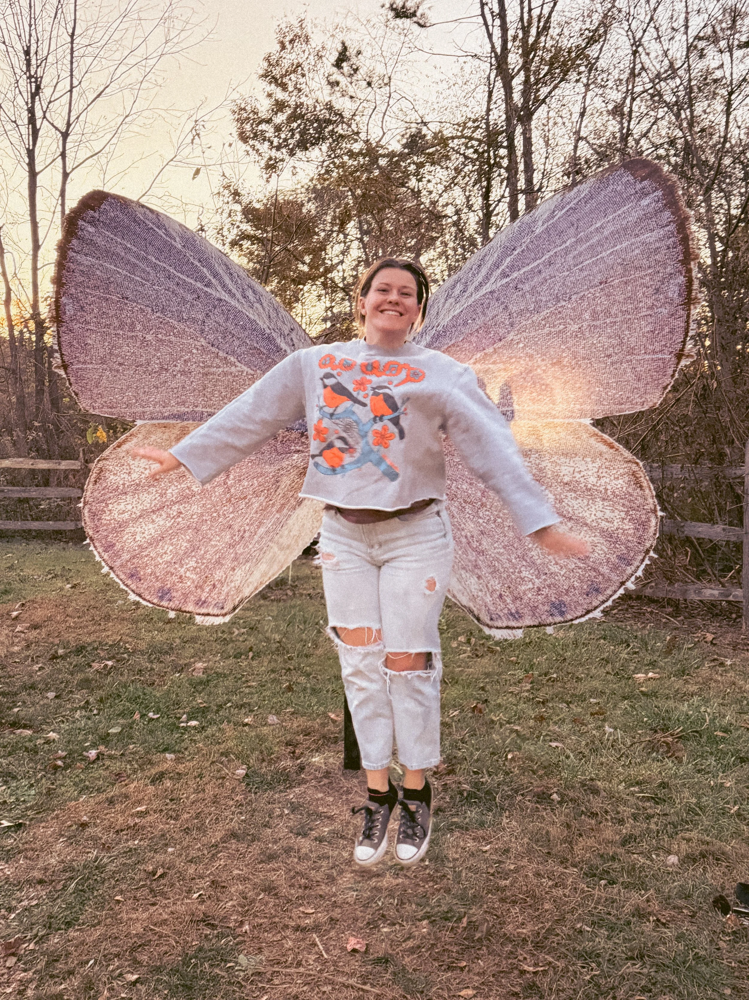 A young woman with short brown hair smiling and jumping outdoors at sunset, wearing a white sweatshirt with colorful bird and floral designs, ripped white jeans, and sneakers, with purple butterfly wings attached to her back.