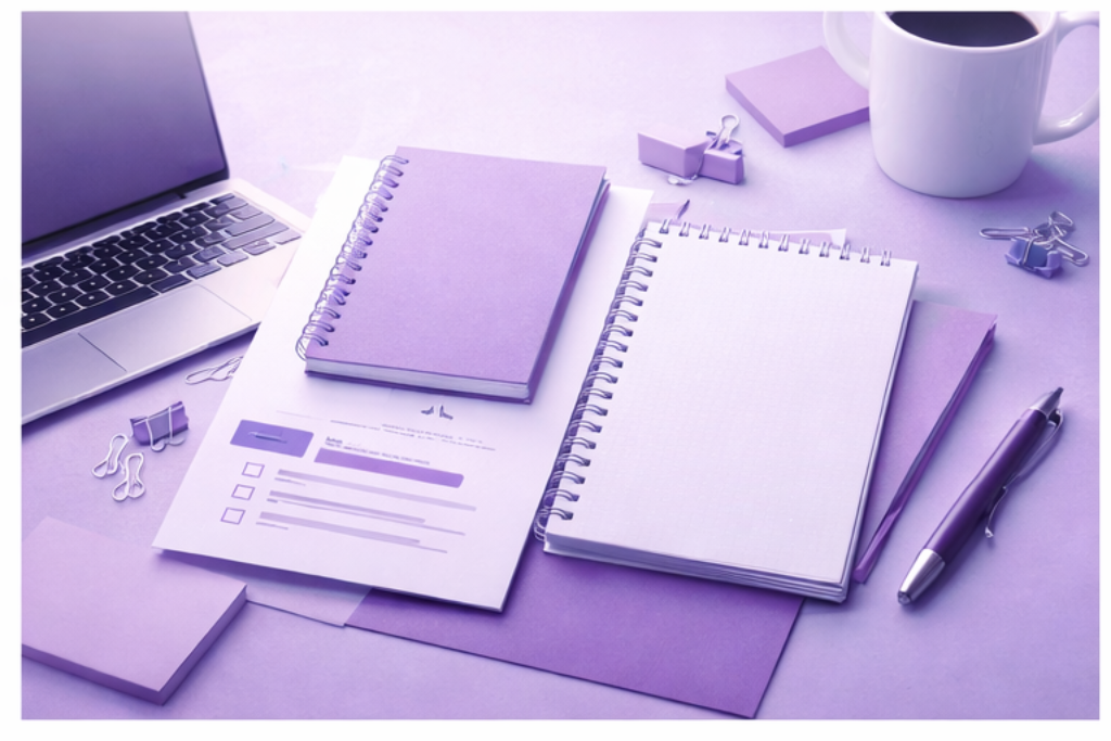 Desk with open laptop, notebooks, a pen, paper documents, sticky notes, paper clips, and a coffee mug, all in purple tones.
