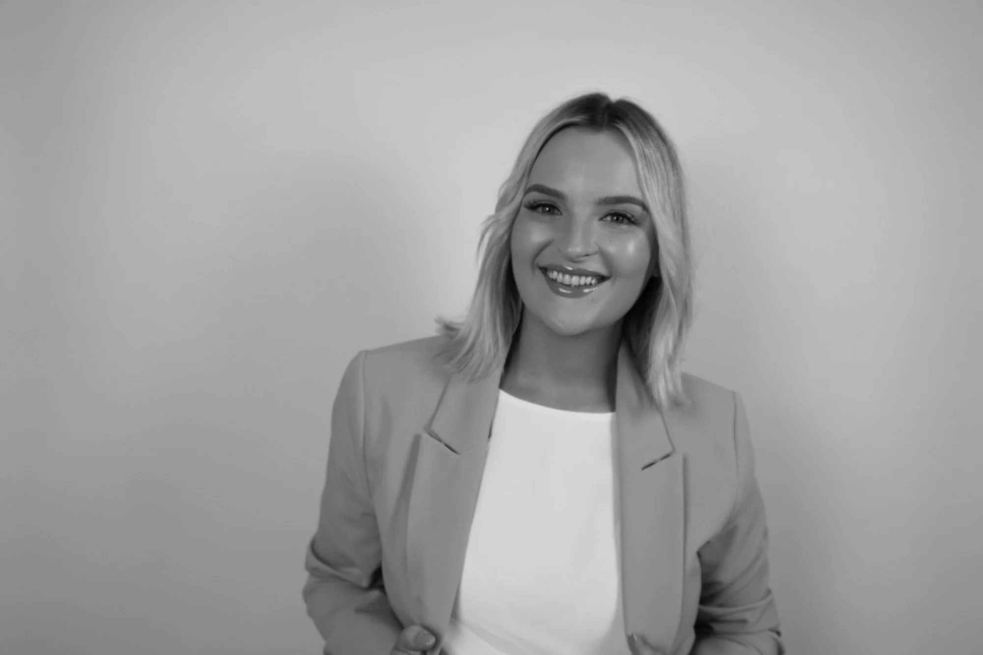 Black and white image of a young woman smiling, wearing a light-colored blazer over a white top, standing against a plain background.