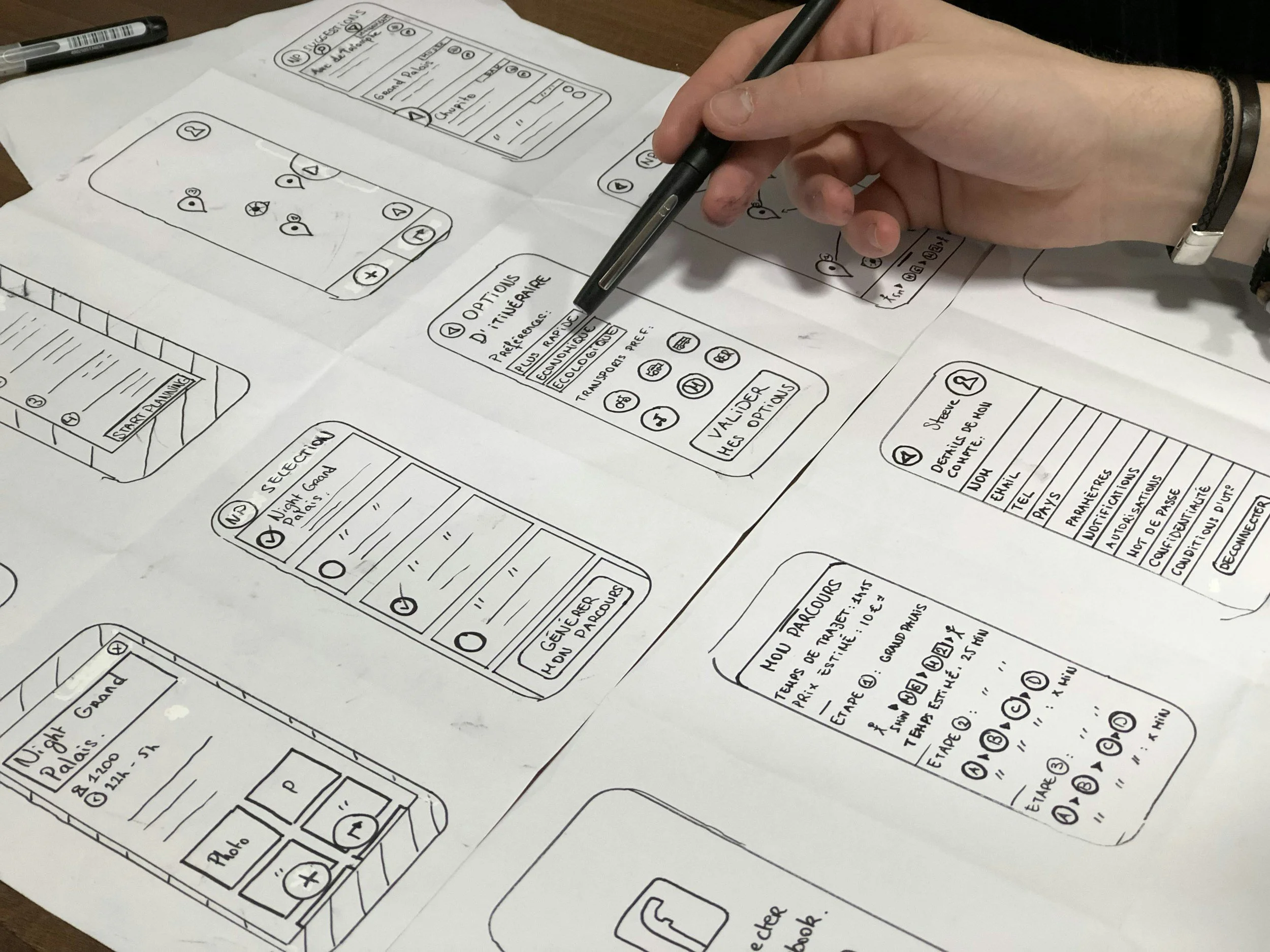 Sketches of a mobile app interface design on paper, showing screens for navigation, options, contact information, and route details, with a person hand-drawing on one of the sketches.
