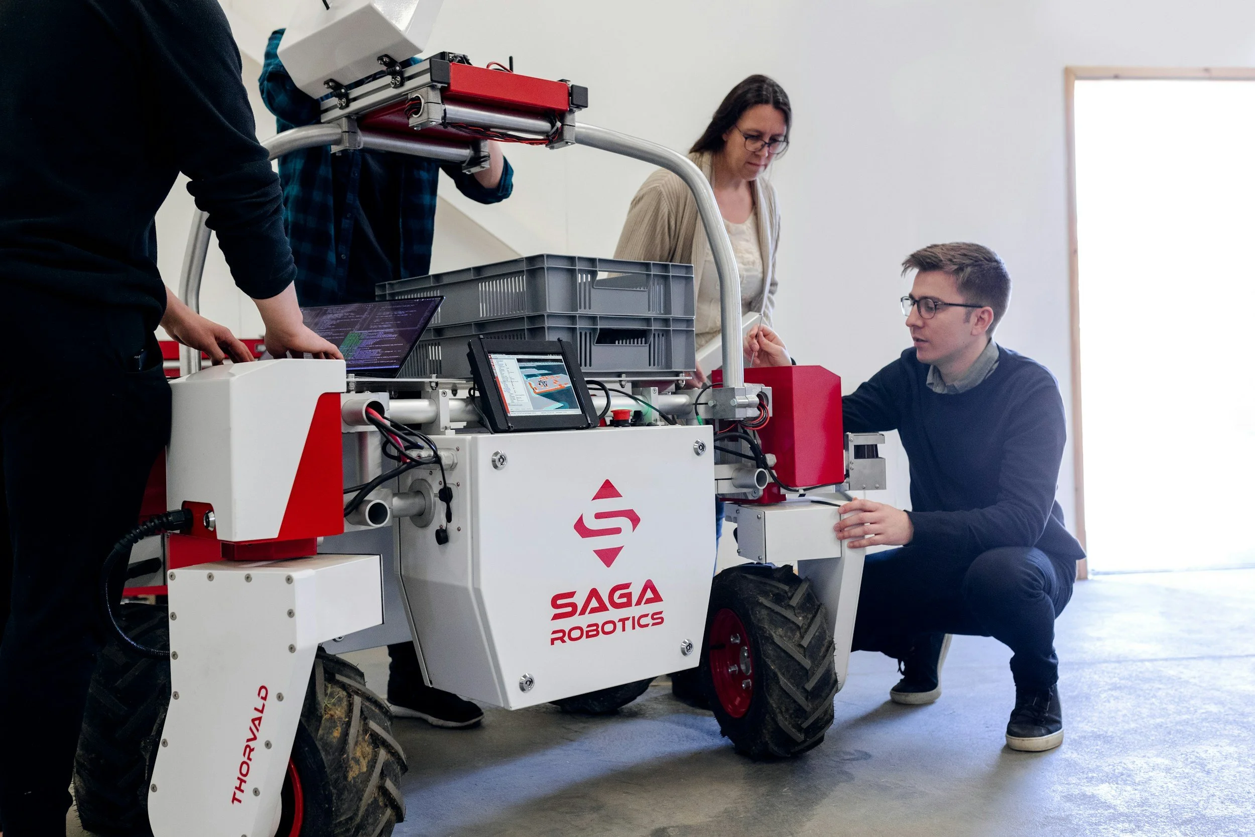 Four people are working on a robotic vehicle labeled 'SAGA ROBOTICS.' One person is kneeling and adjusting a component, another person is standing and observing, and two others are standing nearby, one working on a laptop and the other looking at the robot. The setting appears to be a workshop or laboratory.