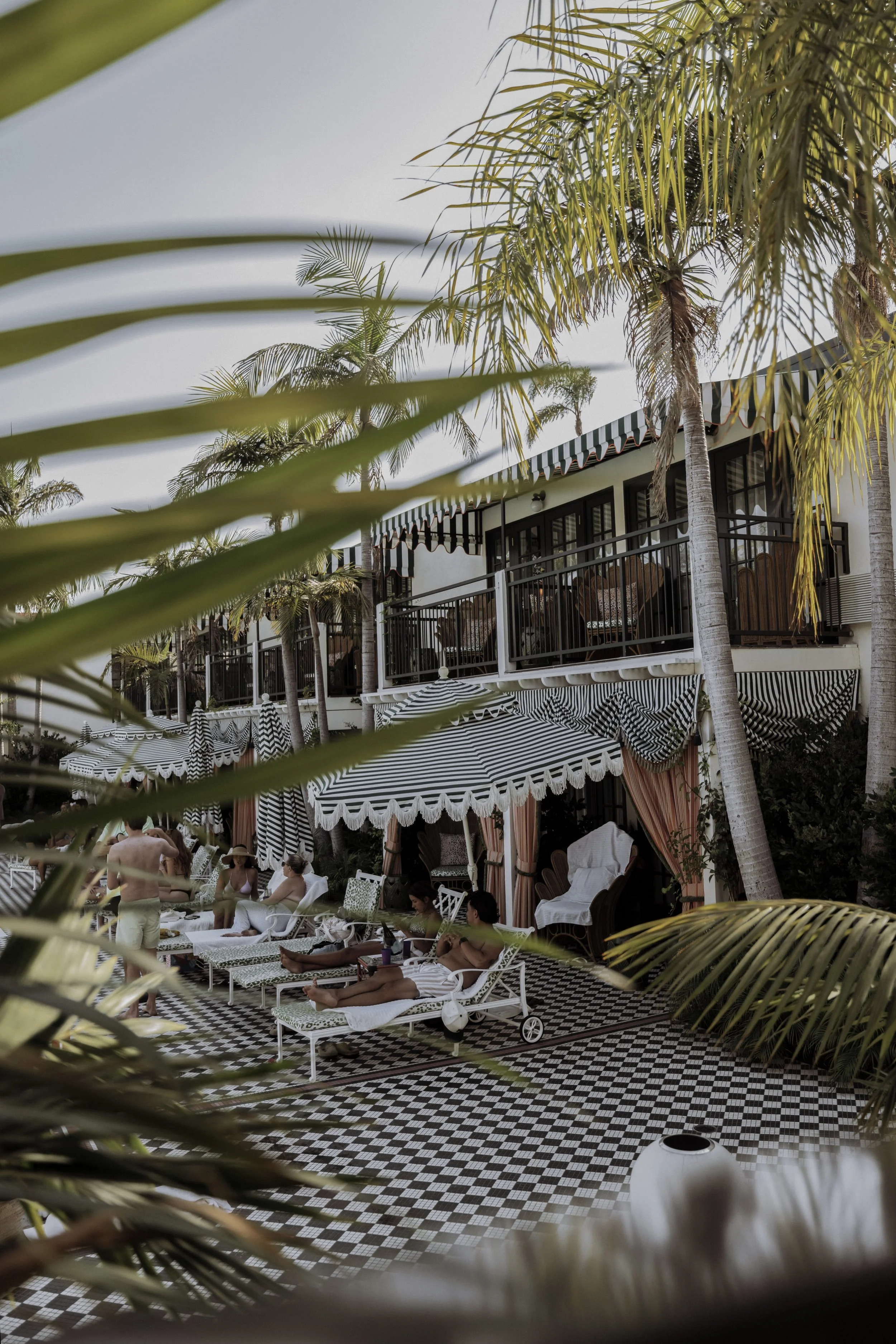 People relaxing on lounge chairs at a poolside patio with striped umbrellas and palm trees.