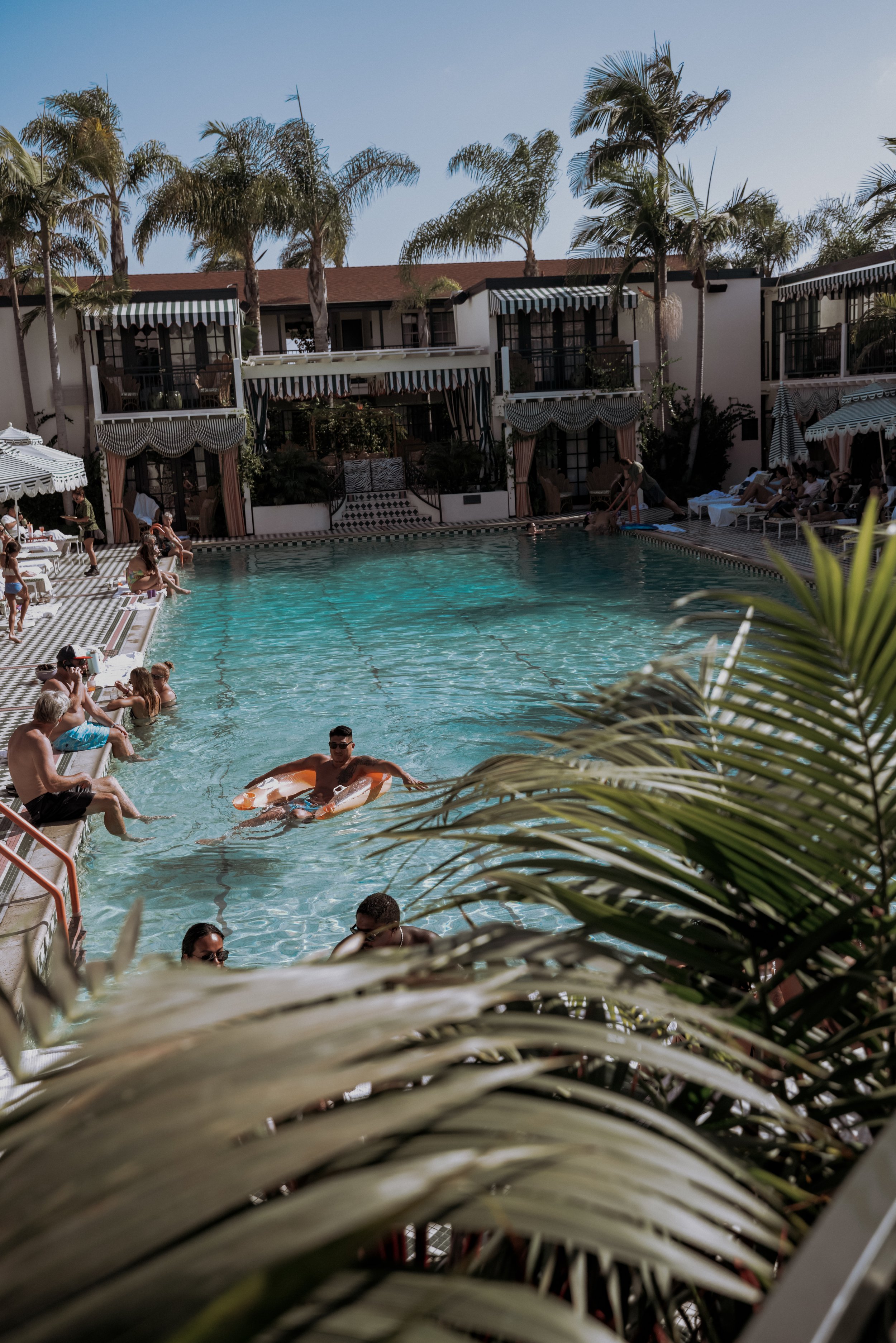 People relaxing and swimming in a sunny resort pool with tropical palm trees and hotel balconies in the background.