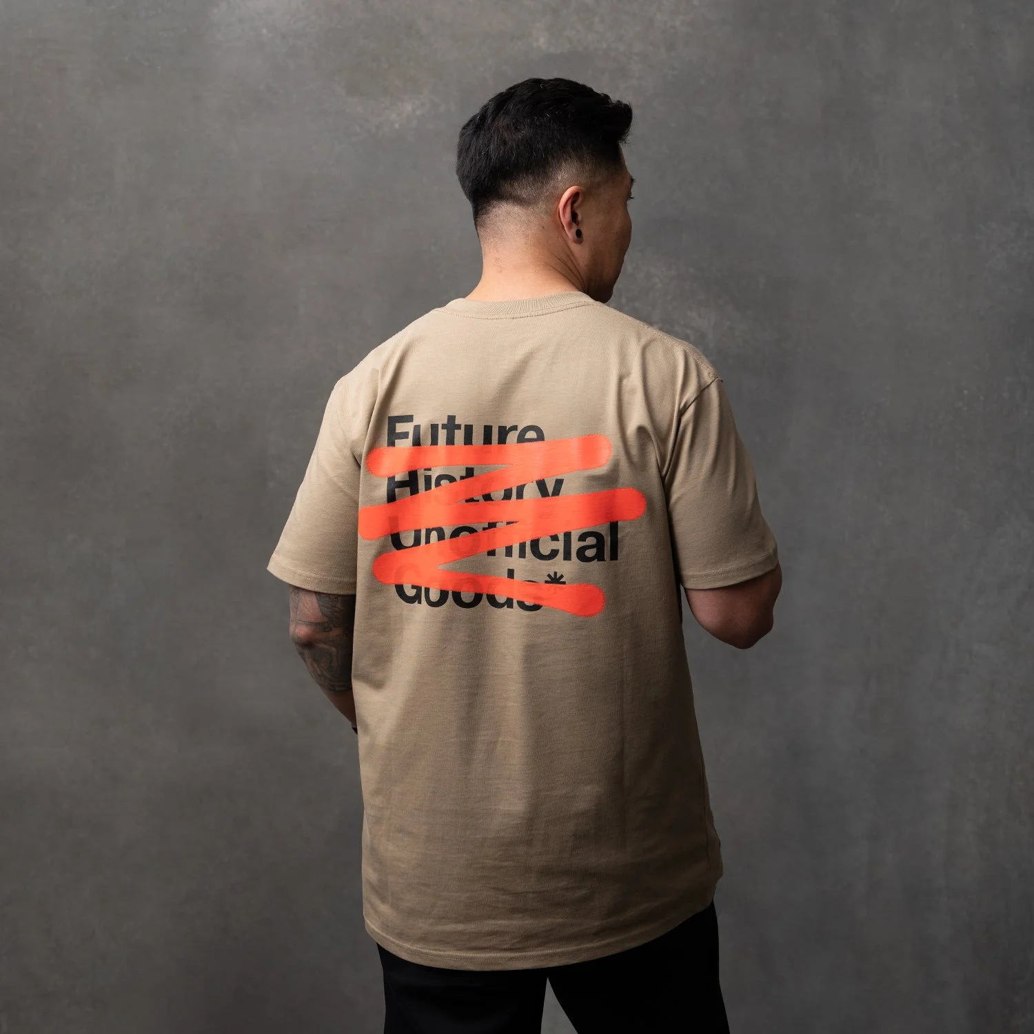 A person with short dark hair and a tattoo on their left arm is standing with their back to the camera against a plain gray background. They are wearing a beige T-shirt with words printed on the back, partially covered by orange brush strokes.