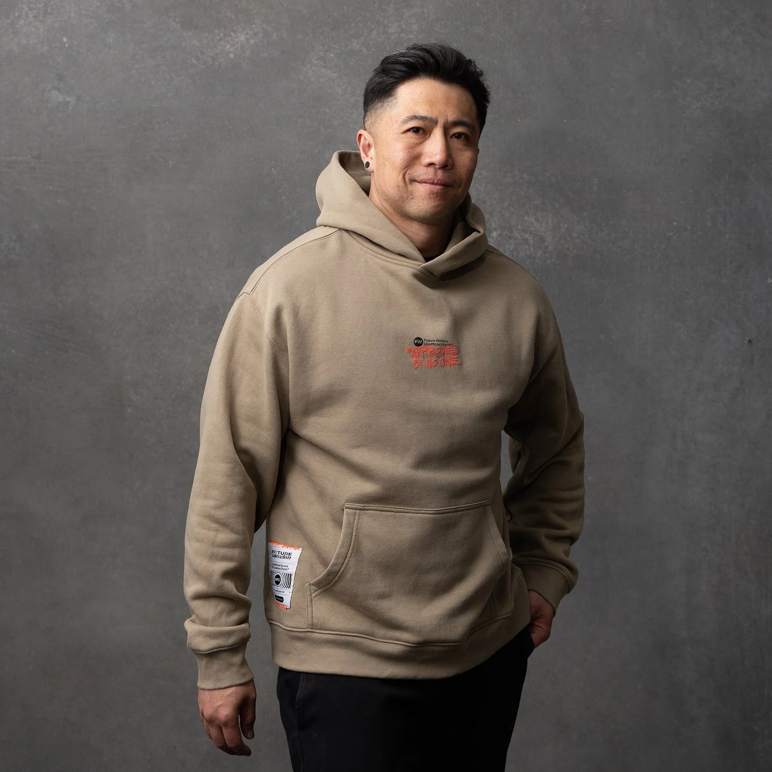 A man wearing a beige hoodie with red and black text and patches, standing against a gray textured background.