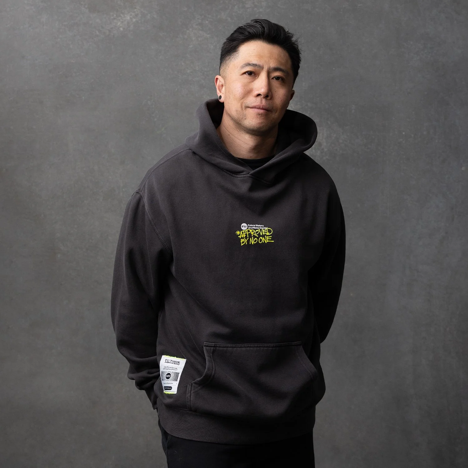 A man with black hair, wearing a black hoodie with yellow text and design, posing against a gray wall.