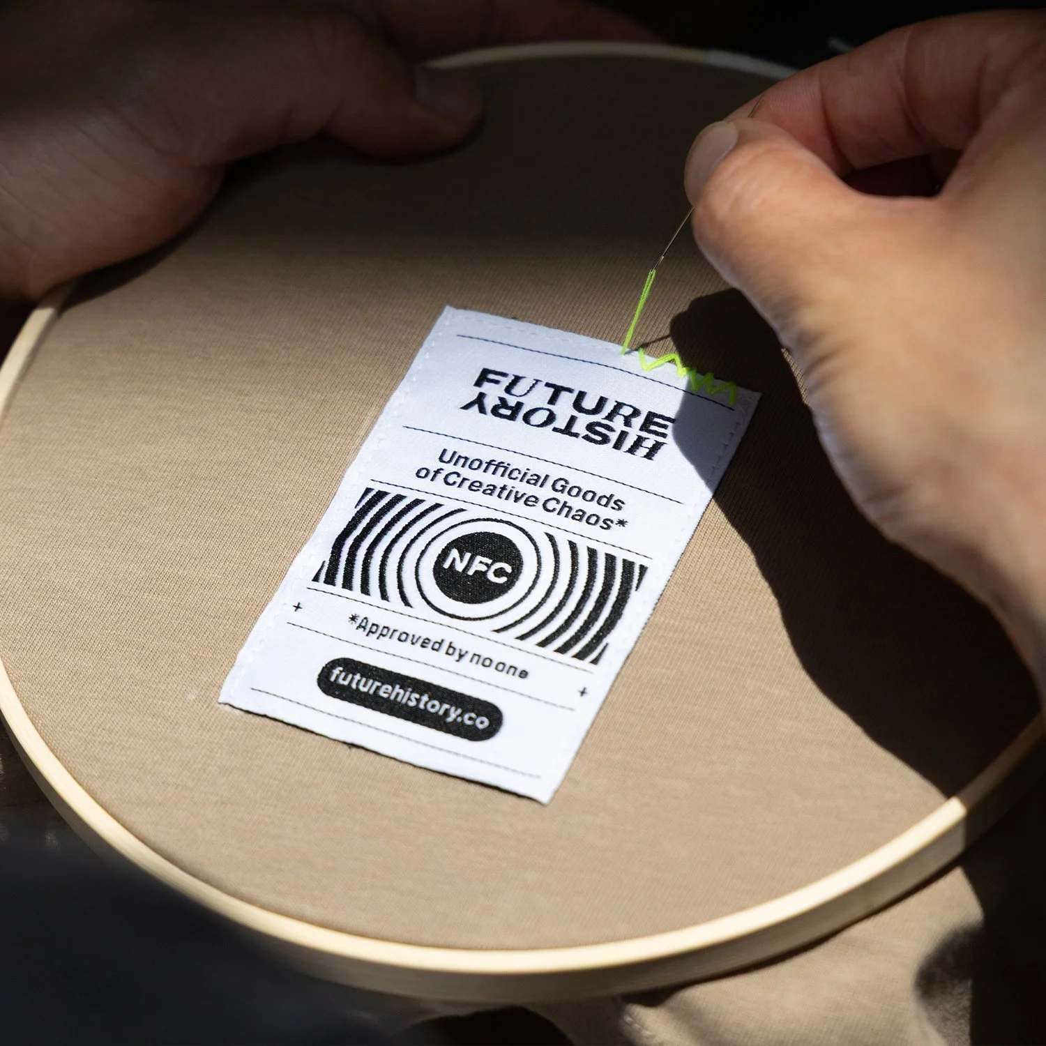 Someone sewing a label onto a beige fabric surface, with the label reading 'FUTURE HISTORY' and 'Unofficial Goods of Creative Chaos,' featuring an NFC symbol and website 'futurehistory.co'.