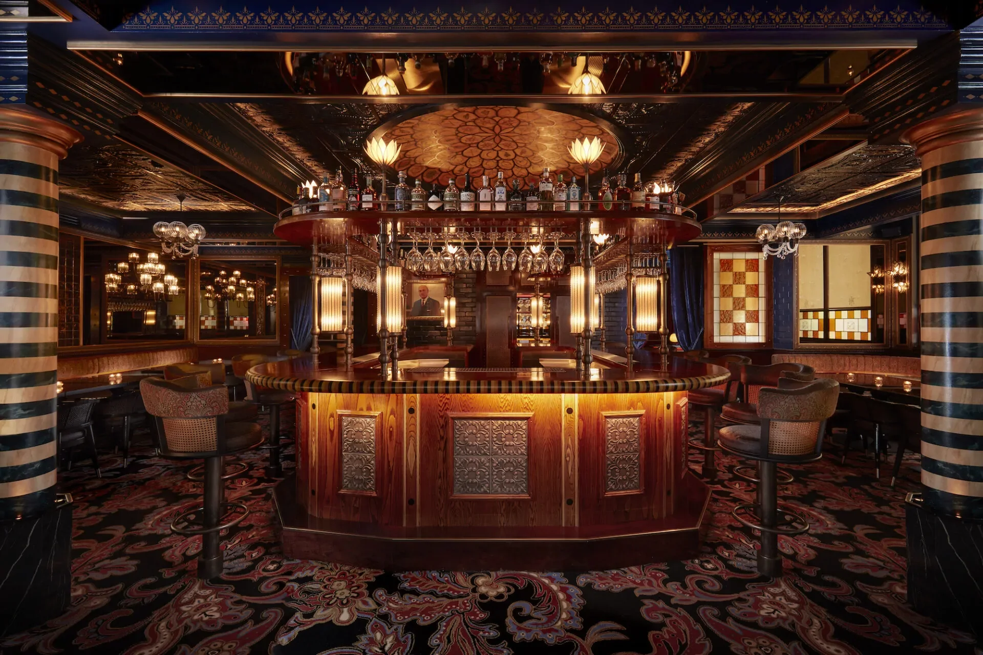 An elegant bar interior with a circular wooden bar counter, glassware hanging above, and bottles of alcohol on a suspended shelf. The room features ornate ceiling detailing, striped columns, and plush seating, with warm lighting and patterned carpet.