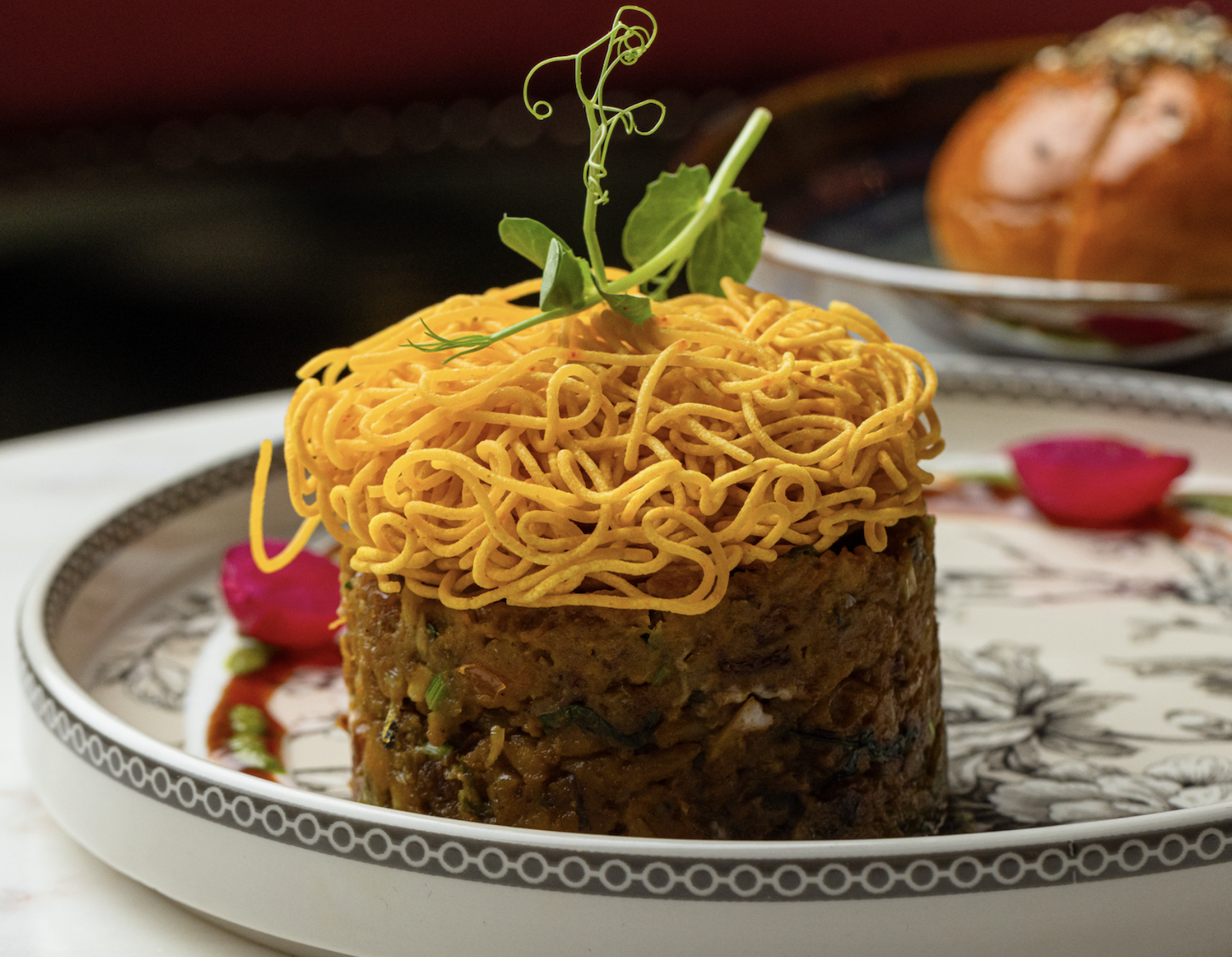 desi food reviews - a rack of noodles garnish on top of a mound of minced eggplant with herbs, served on a decorative plate with flower petals. In the background, there is a bowl of bread roll.