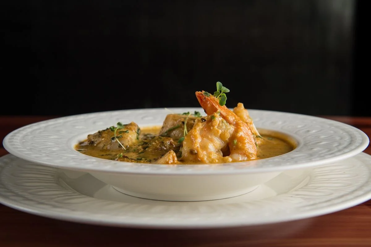 A white plate of seafood curry with shrimp, fish, and herbs