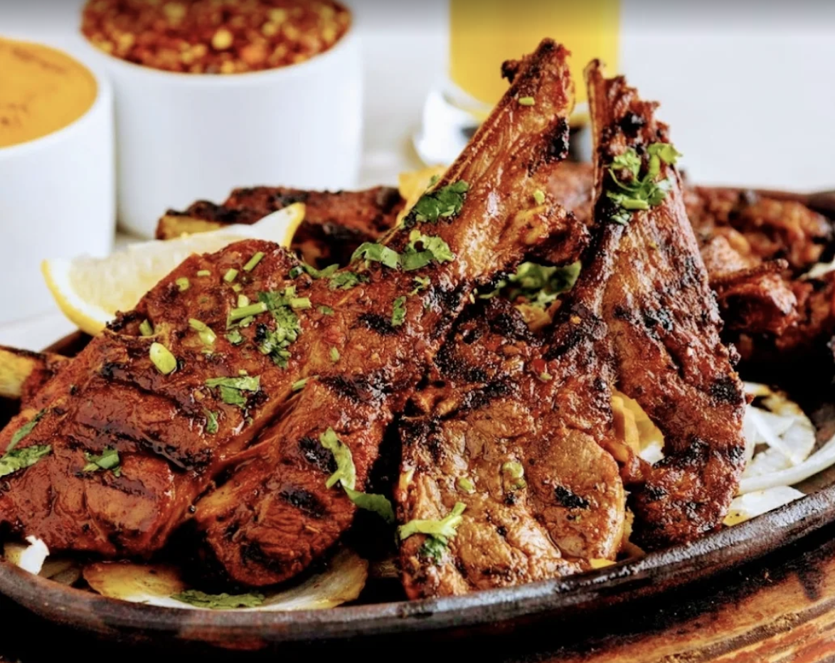 Close-up of Aga's world famous lamb chops grilled in a tandoor, garnished with cilantro and lemon wedges, served on a platter with additional side dishes in small bowls in the background.