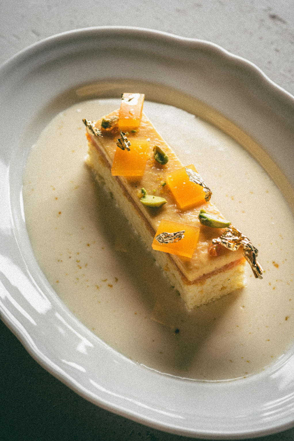 A slice of layered cake garnished with diced fruit, pistachios, and edible gold leaf