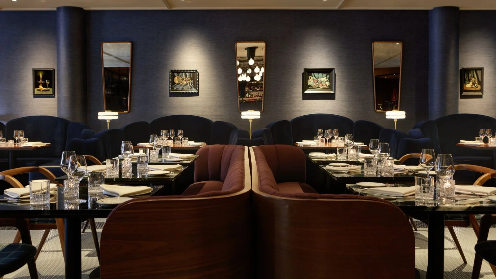 Elegant restaurant dining area with tables set for service, featuring wine glasses, water glasses, and napkins, with modern artwork and mirrors on the dark blue walls.