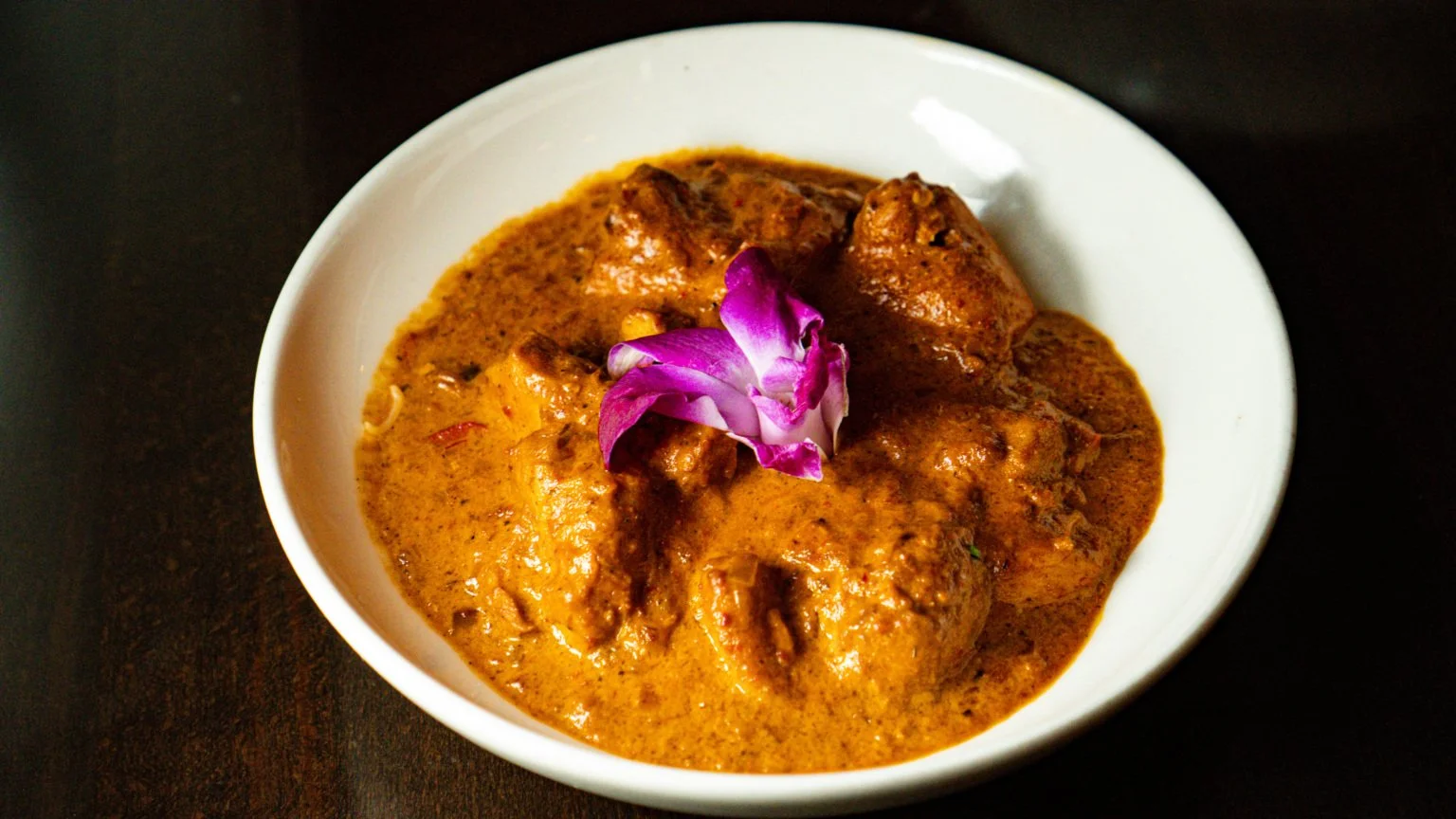A white bowl of Indian chicken curry garnished with a pink flower