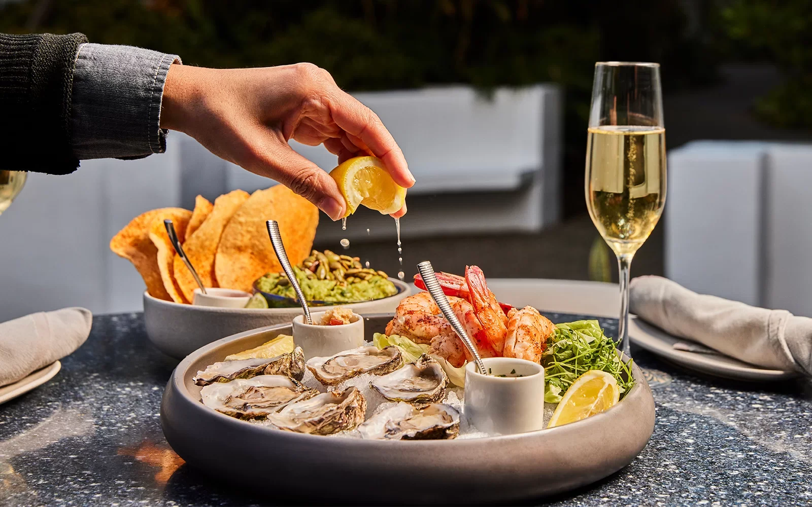 Person squeezing lemon over a platter of oysters, shrimp, and condiments on a dark table.